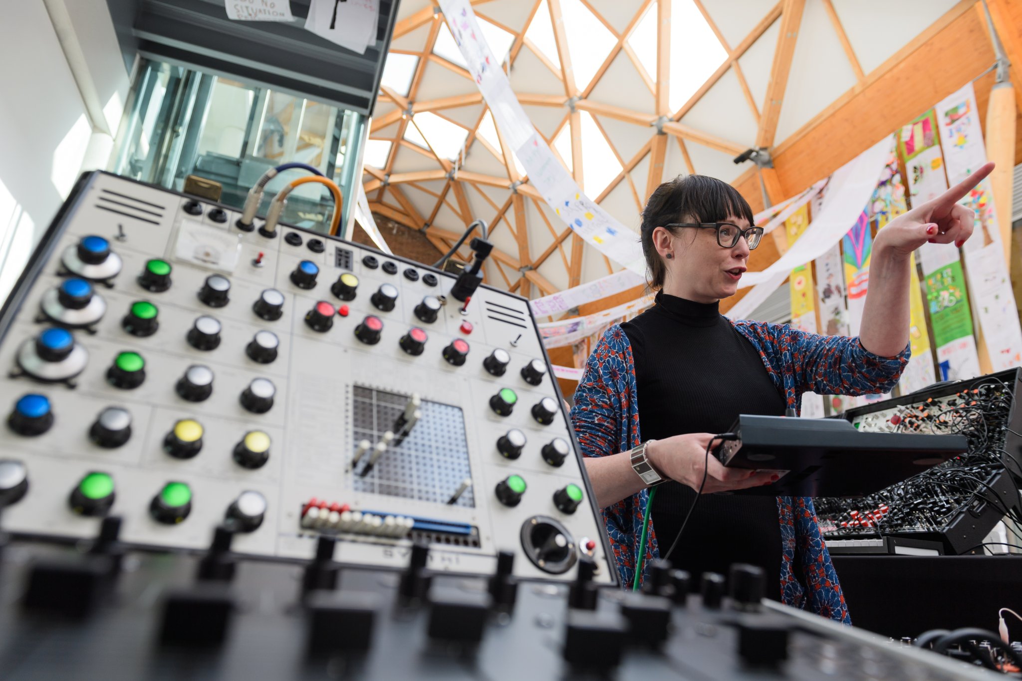 A woman holding a keyboard and pointing, standing next to a synthesiser in the Herbert's covered court