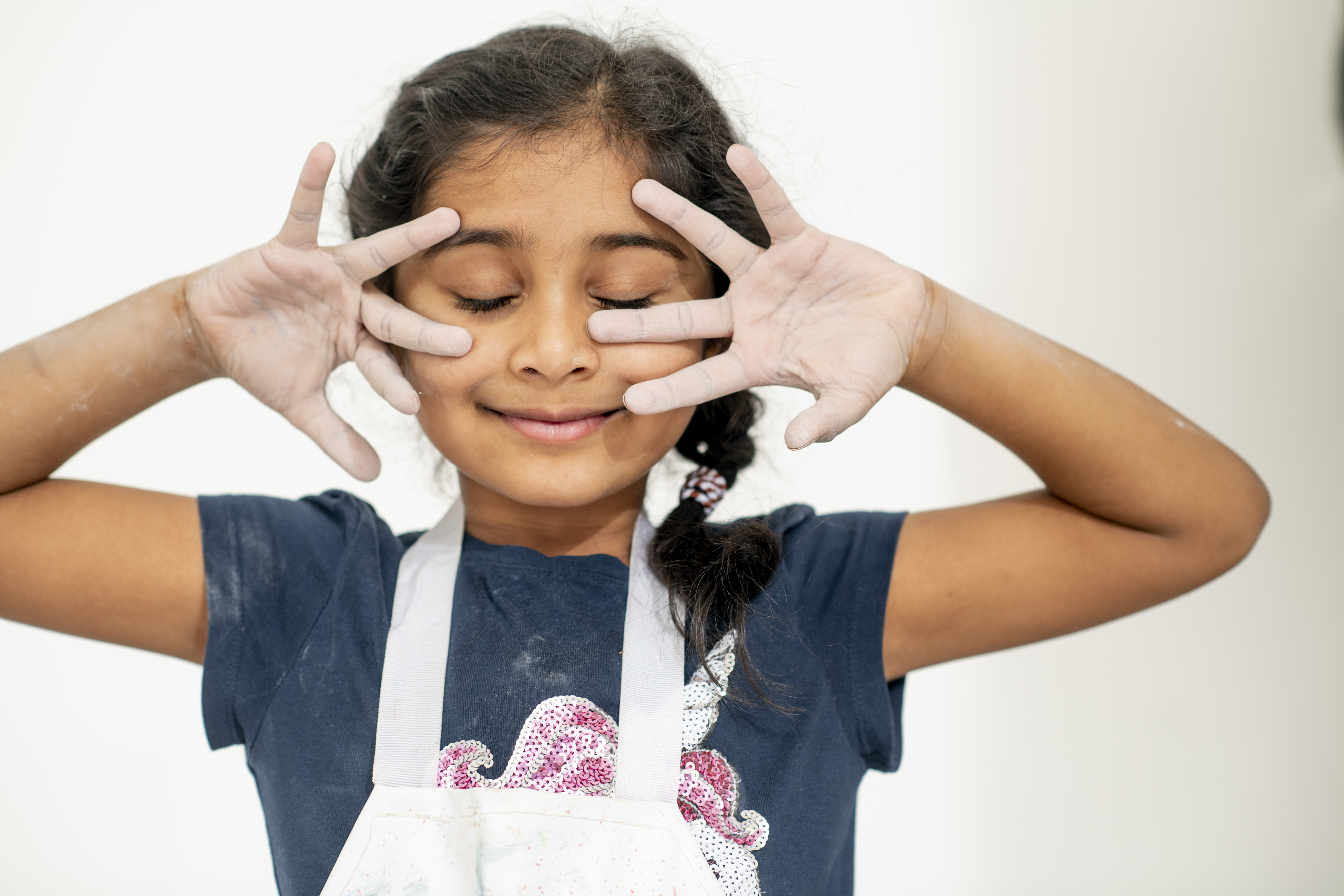 A girl with plaited hair wearing a white apron holds her hands up to her face, showing us her palms, which are whitened with clay dust