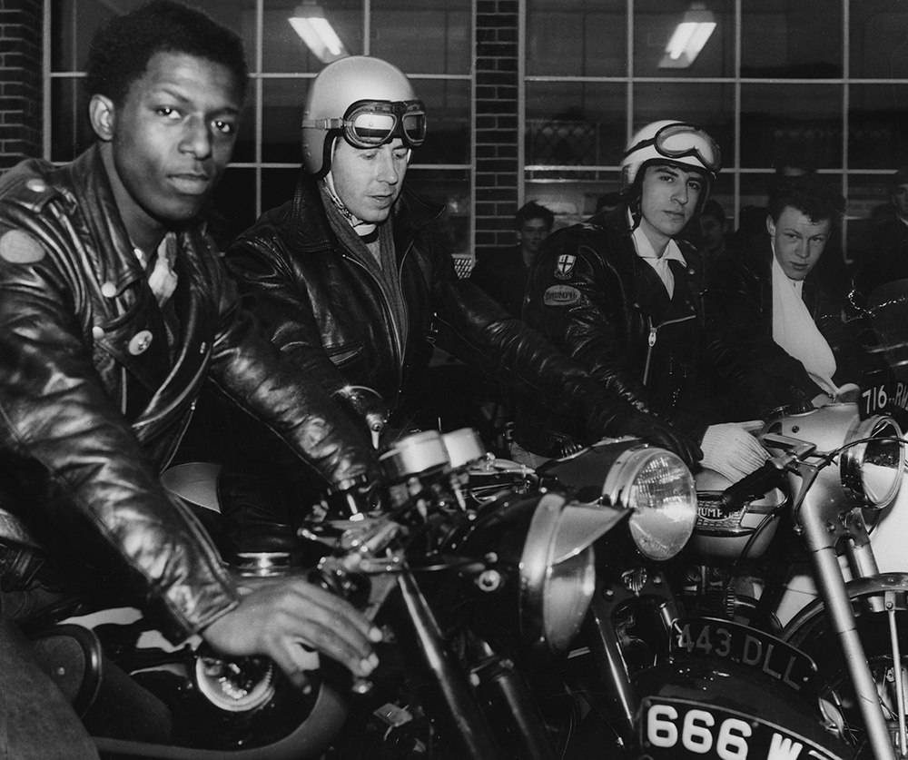 A black and white photo of a group of young men in black leather jackets, sitting on motorbikes outside a building