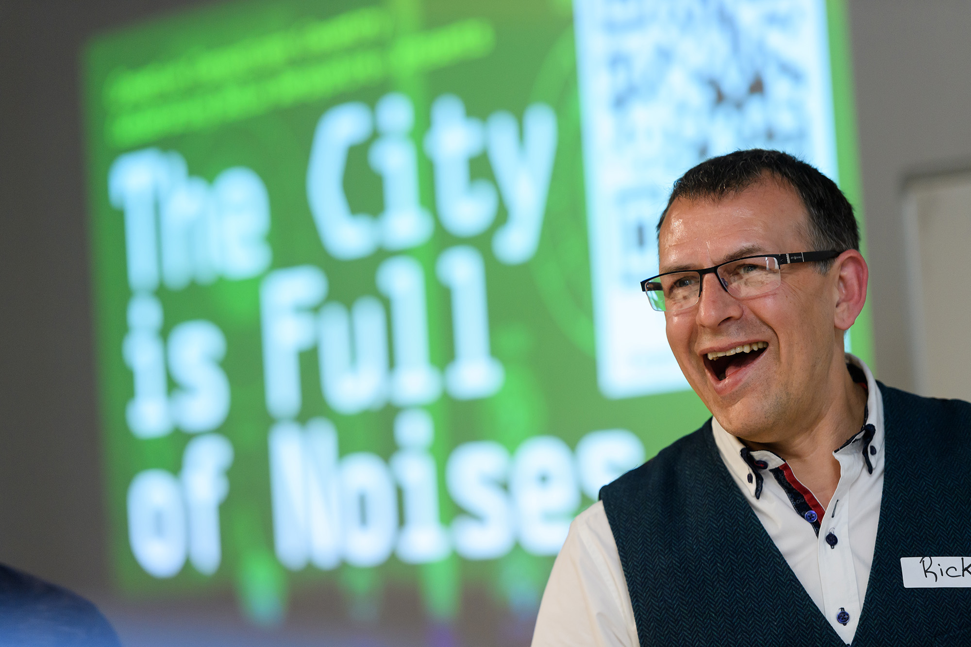 A photo of Rick Holt from Frequency Central smiling. He is standing in front of a screen with "The City is Full of Noises" projected onto it
