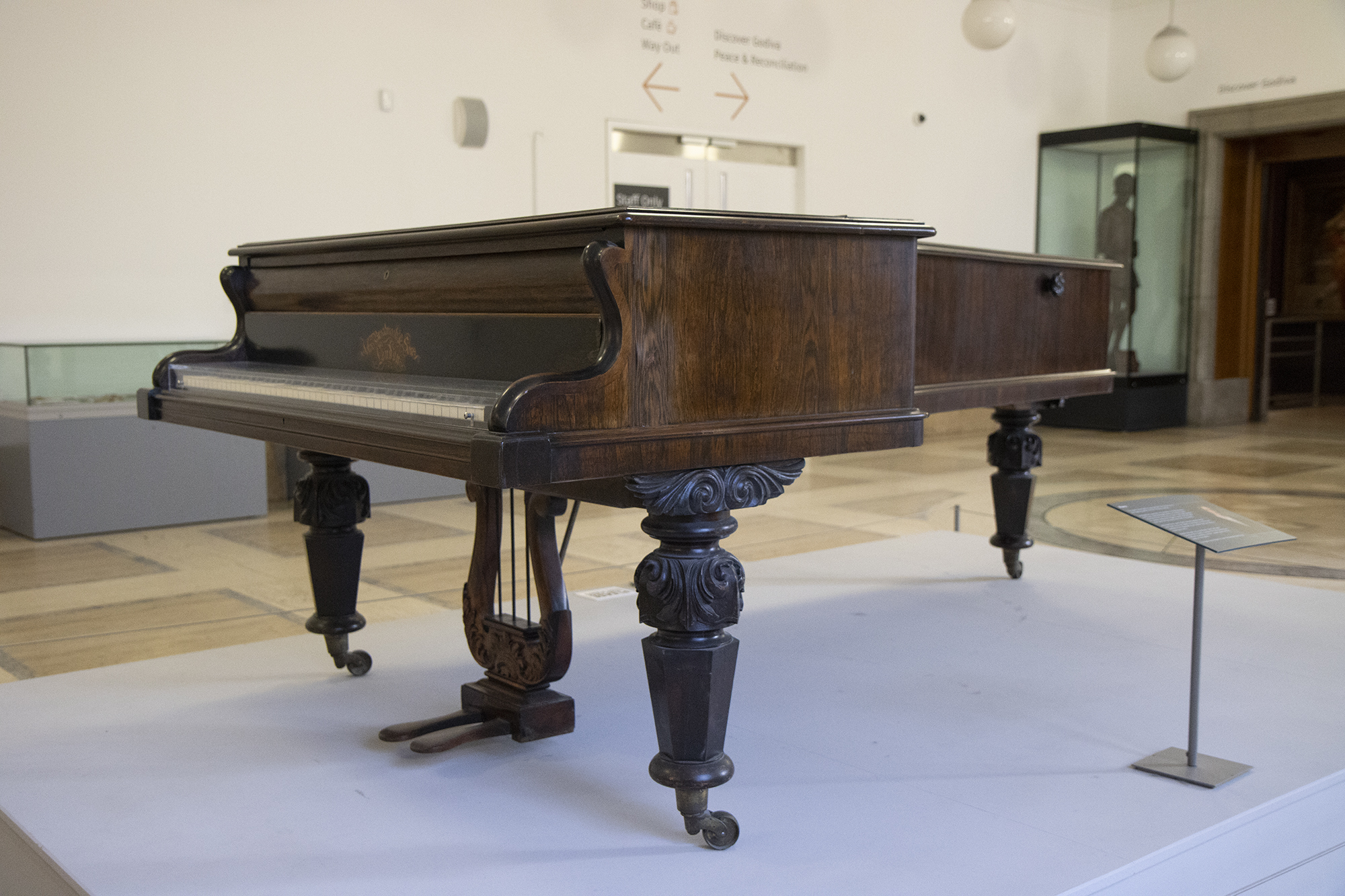 A photograph of George Eliot's grand piano, on display in the Herbert Art Gallery & Museum foyer. It's made from dark wood with elegant swirled carvings on the legs. The pedals are attached to a harp-like carved design underneath the piano. Above the keys is the manufacturer's name.