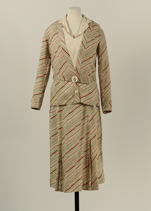 A woman's beige 2-piece skirt suit a colourful chevron striped pattern in pink, turquoise and brown. The outfit is matched with a cream blouse and the jacket features a matching belt with a decorative buckle