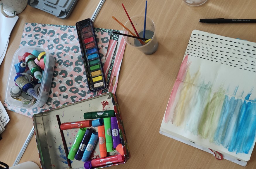 An array of watercolour paints, coloured markers, stamps and glitter glue laid out on a table next to an open journal, with a rainbow design painted on the pages