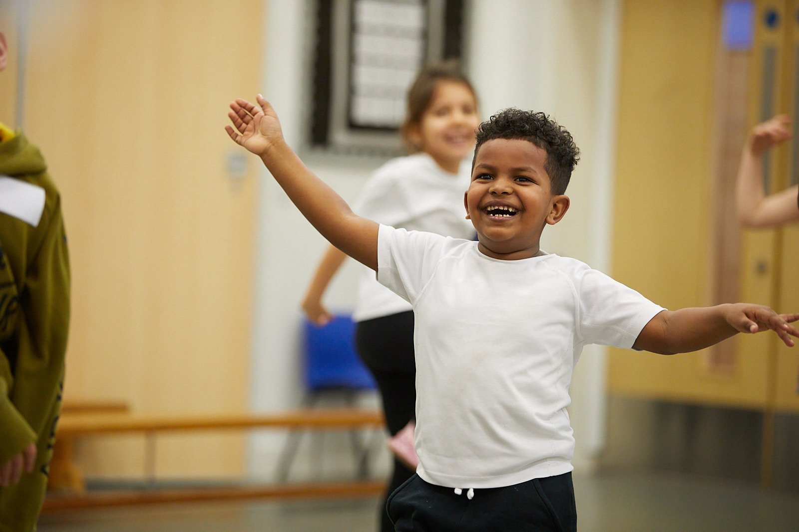 A laughing boy faces the camera with outstretched arms. In the background a girl is following him.