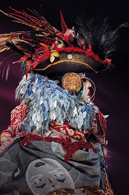 Daniel Lismore sculpture in profile featuring a feathered blue scarf, gold disc-shaped earrings and an 18th century-style hat loaded with red and black feathers