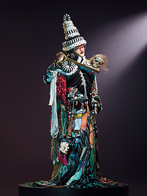 Daniel Lismore sculptural ensemble featuring multicoloured skirts, a layered hat with black and white stripes and spots, a Betty Boop handbag and a skull perched on the shoulder.
