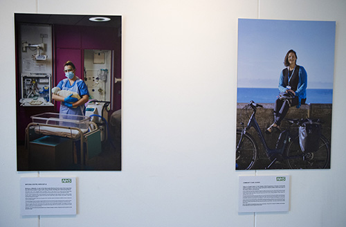 Two photo portraits of NHS staff. On the left, a woman in blue scrubs holds a baby next to a cot on a maternity ward. On the right, a woman in a blue cardigan sits on a wall with the sea behind her and a bicycle resting against the wall in front of her