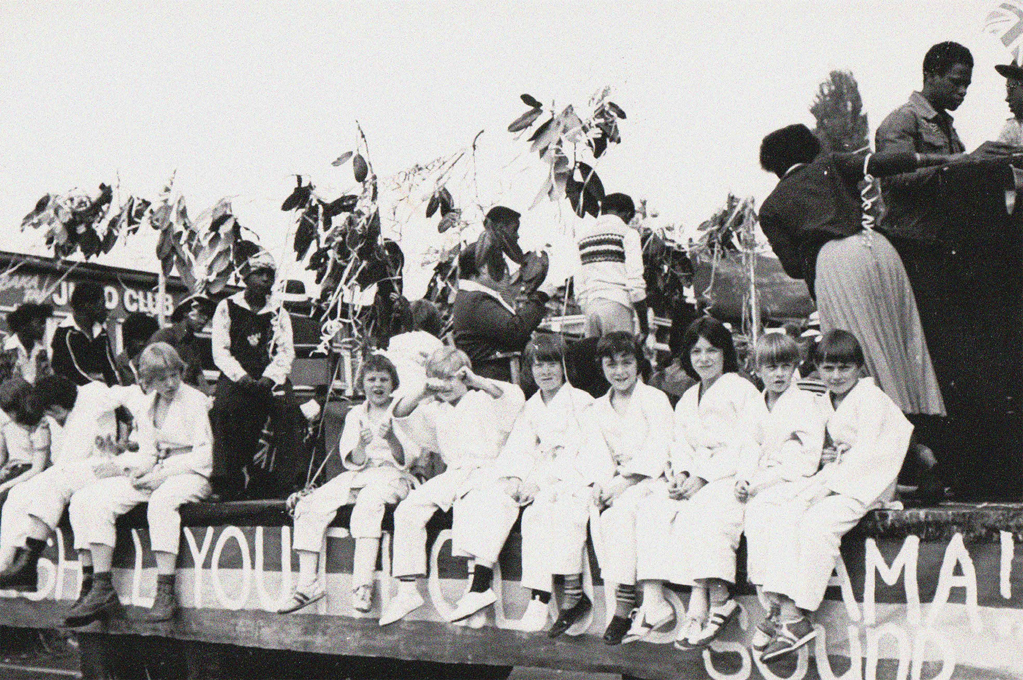 A black and white photograph of a stage with text painted on the side reading, "Foleshill Youth Club Jamaican Sound". There are Black adults and children on the stage setting up, presumably for a concert or party, while a group of white children sits on the edge of the stage, posing for the camera.