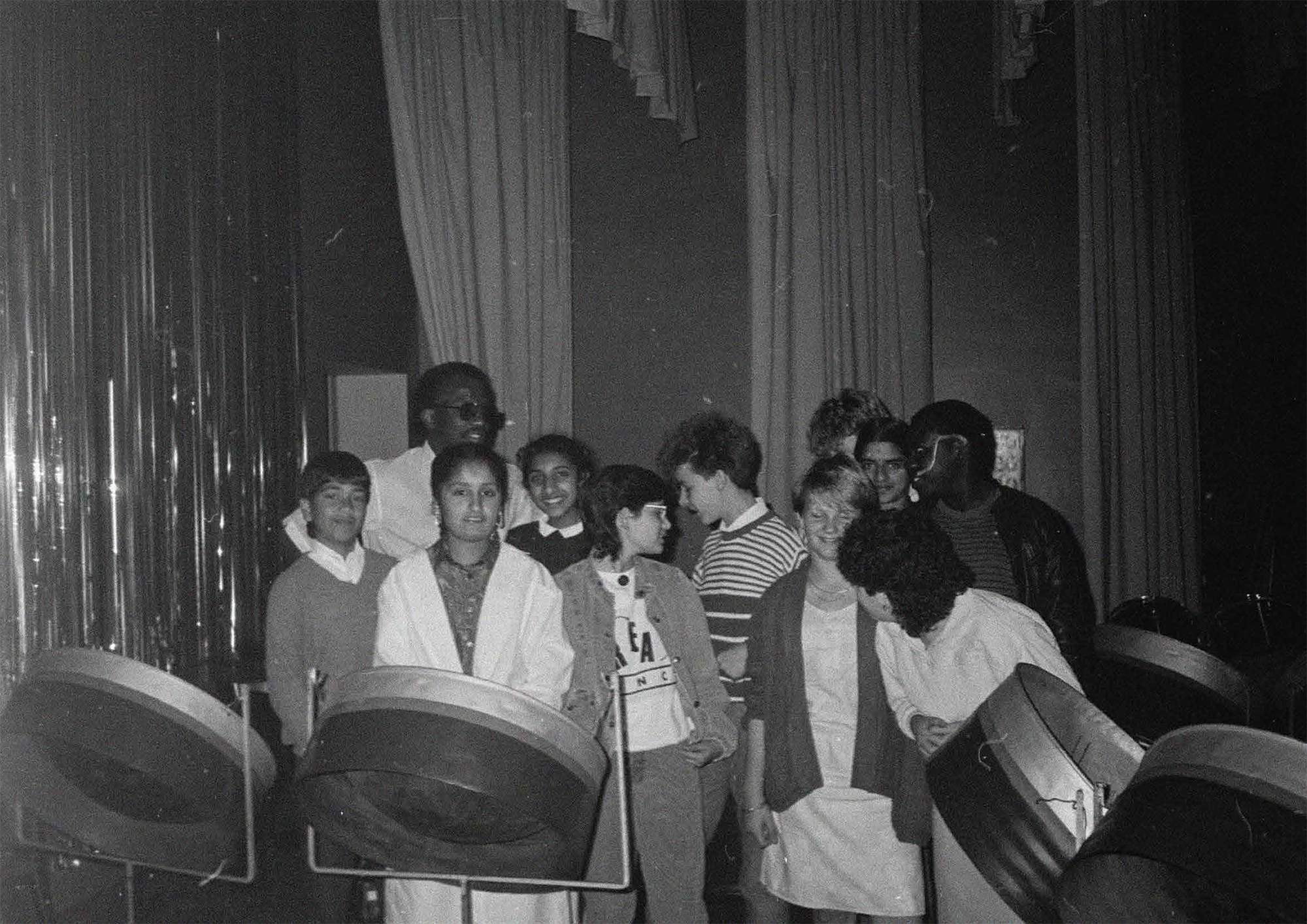 A group of teenagers of different races standing together in the midst of a set of steel drums