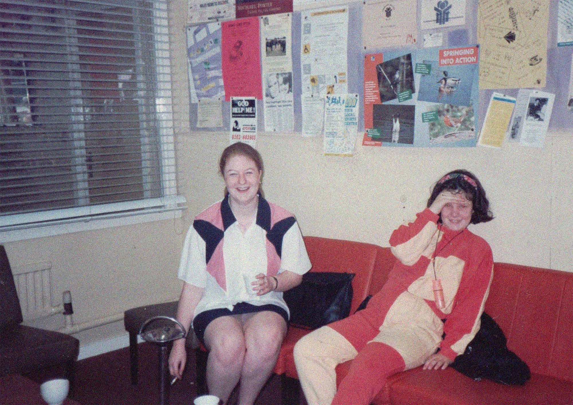 Two older girls or young women sitting on chairs under a notice board, smiling towards the camera. They are wearing bright 80s or early 90s style block colour clothing.