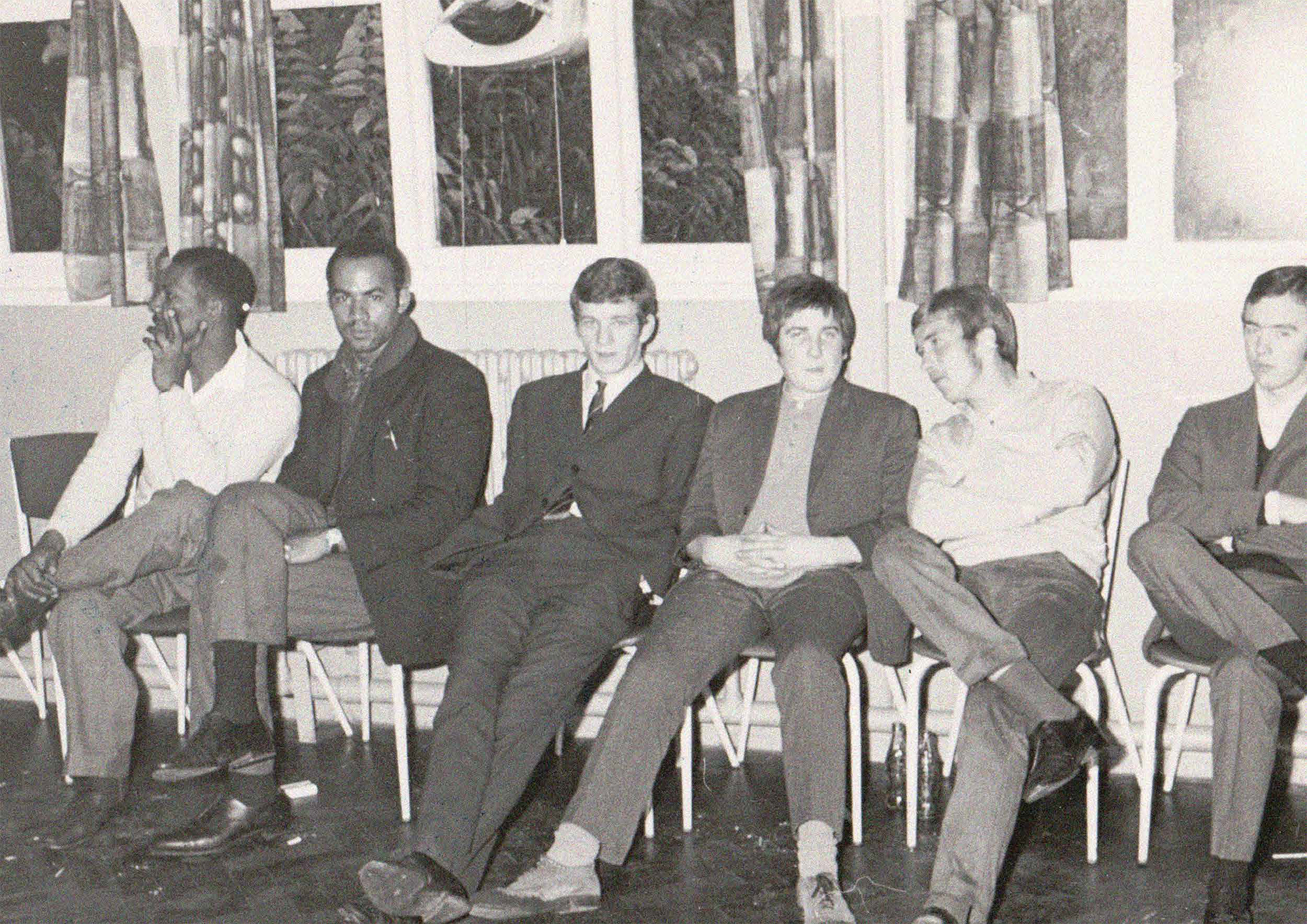 A group of young men sitting on chairs in Foleshill Community Centre's main hall, with heavily patterned curtains and a leaf print on the windows behind them. Some are turned towards the camera and some are chatting with each other.