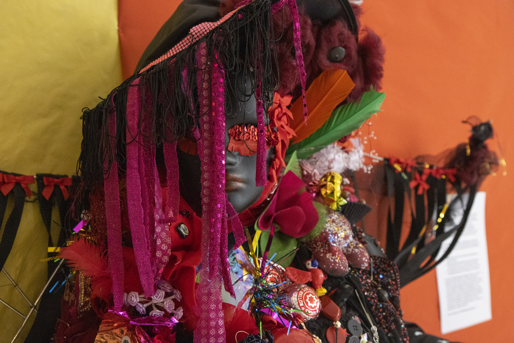 A sculpture made from a black mannequin, dressed in red and black colours including a big fabric had with black tassels and pink glittering ribbons, red sequin eyes and black mesh wings. Feathers, tunnocks teacakes and flower sprays decorate. 