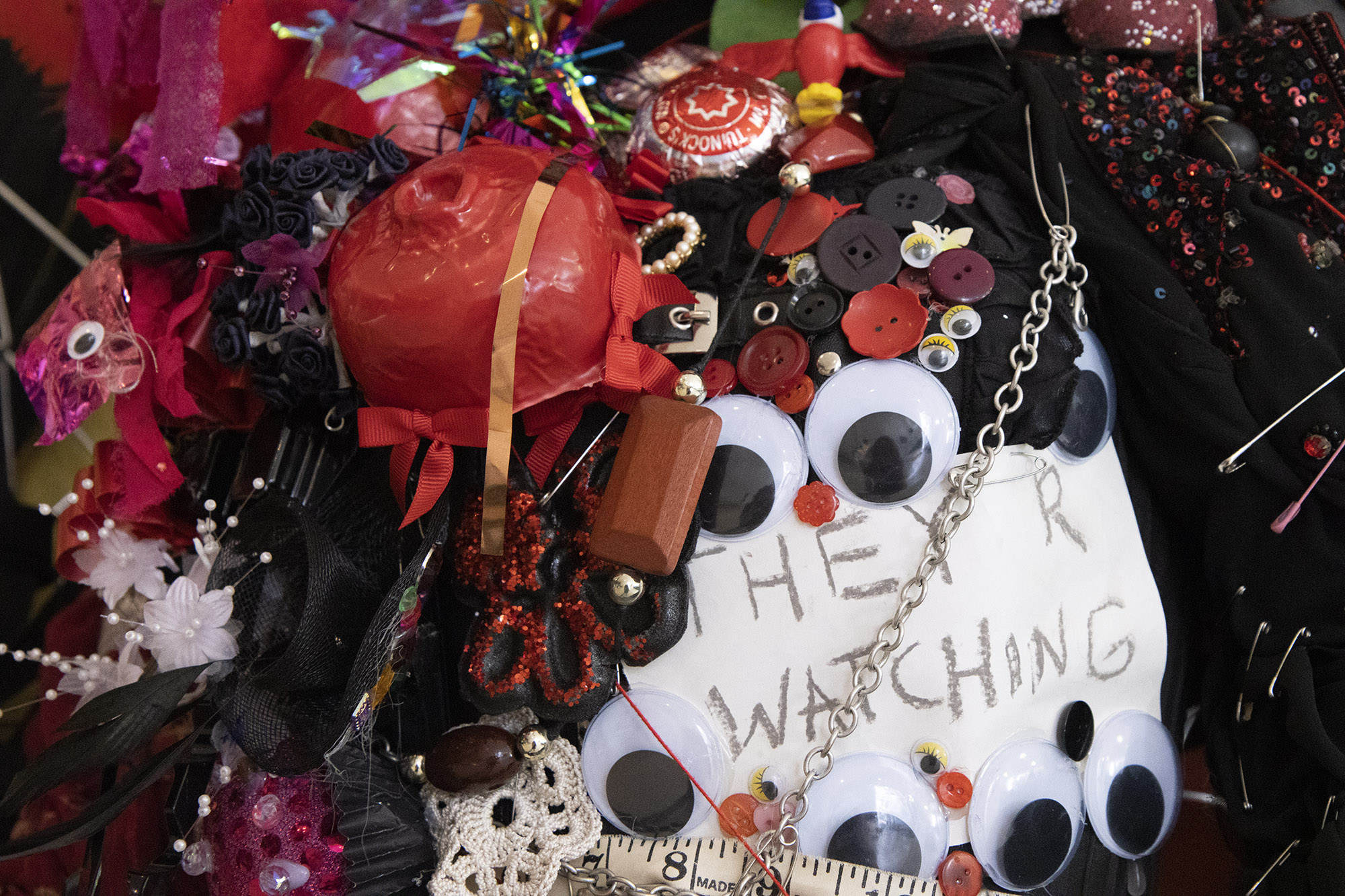 Close-up of a section of a sculpture featuring a vacuum-formed pomegranate, black mesh, flower sprays, tunnocks teacakes, crocheted lace, a dressmakers tape measure, buttons and google eyes around a piece of paper with the text, "THEY R WATCHING"