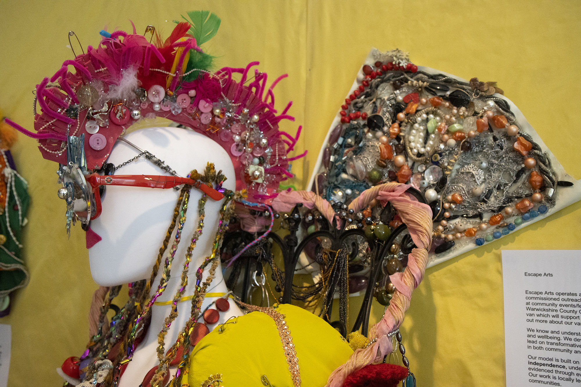 A mannequin sculpture with a pink mohawk made using pipecleaners and buttons. The mannequin has red lips and is wearing red sunglasses with heart-shaped mirrors faced to the front. Behind it are wings covered in found beads and buttons.