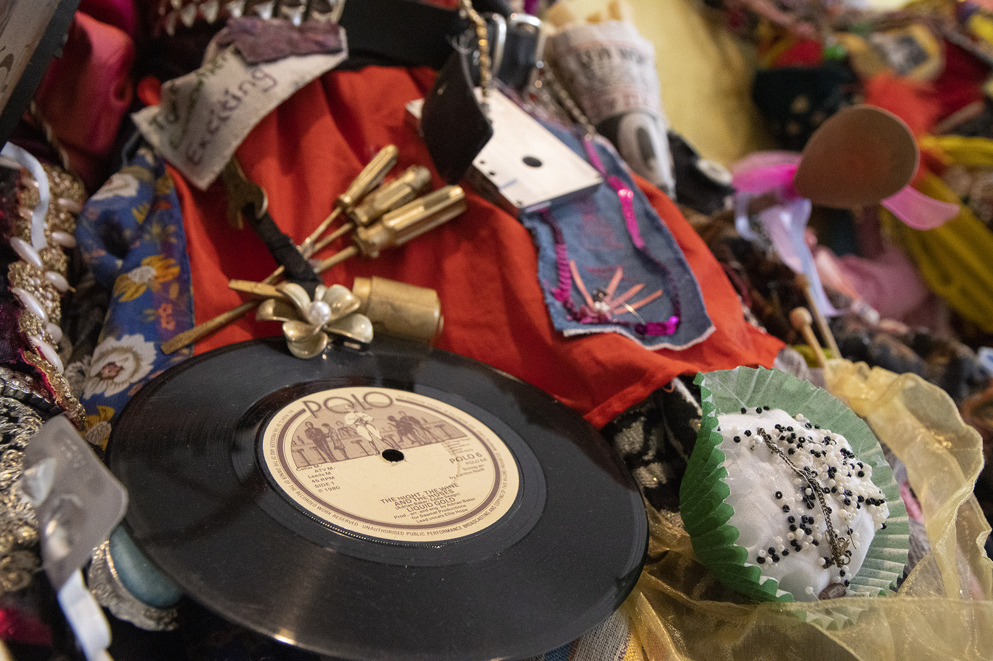 A section of a sculpture featuring a vinyl record, a cassette, gold-painted screwdrivers, a white fabric cup cake in a cupcake case, decorated with black and white seed beads and bits of jewellery and fabric. 