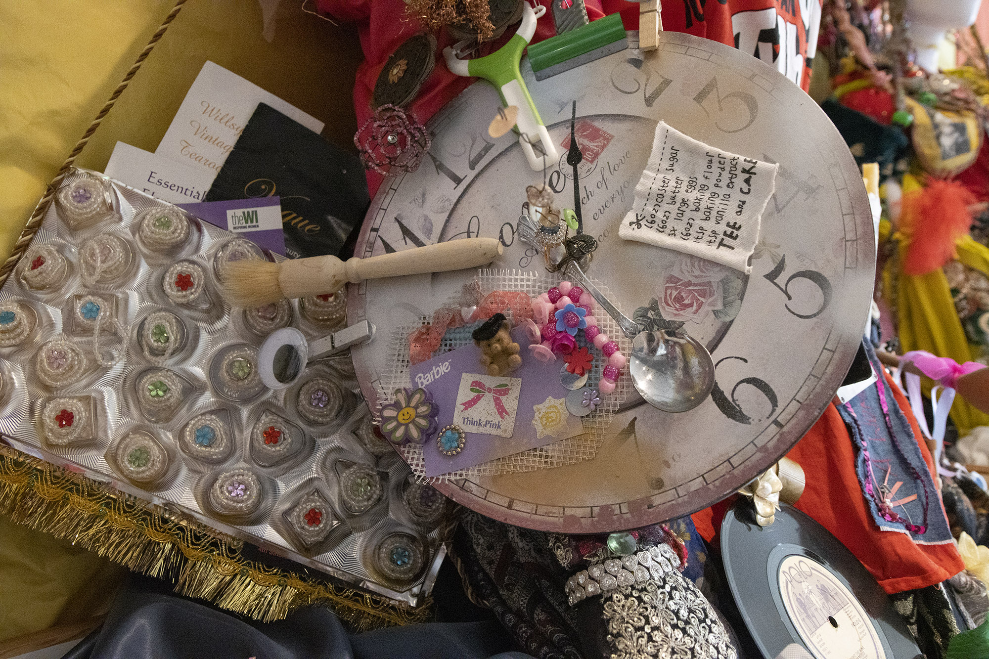 A secret of a sculpture featuring a clock face decorated with objects including a pastry brush, Barbie accessories, a spoon, a vegetable peeler, a clothes peg and a small receipt for cake. Beside it is the plastic tray from a box of chocolates, filled with little beaded shapes.