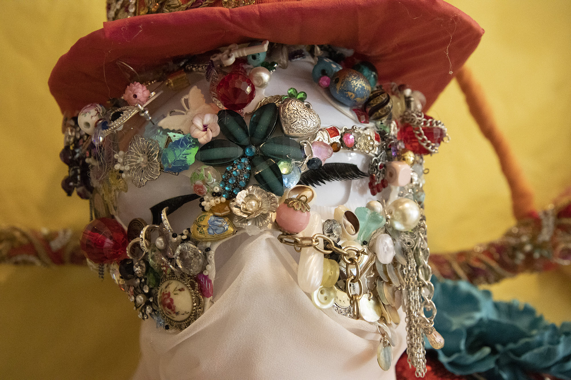 A close up of the face of a white mannequin covered with a mask, decorated with buttons, beads, gems, chains and sequins. False eyelashes stuck to the sculpture are visible in cut-out areas of the mask, and the nose is covered with a light, white fabric.