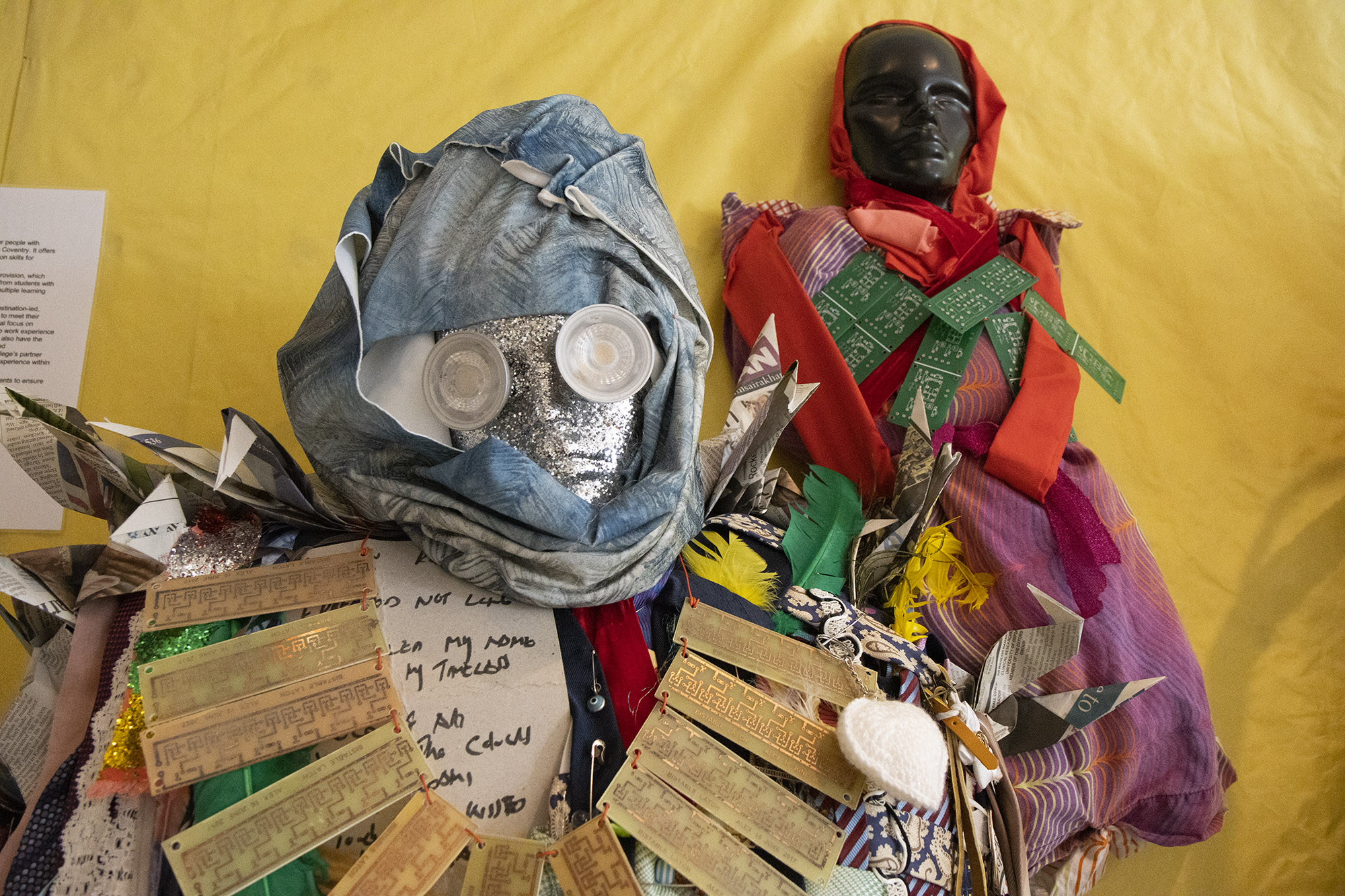 A smaller sculpture hangs on the wall behind a larger one, both with necklaces strung with circuit boards. The mannequin in the foreground has a glittering silver face, plastic lid eyes, a blue fabric head scarf, and origami cranes on its shoulder. The smaller mannequin has a black face with a red headscarf and dress.