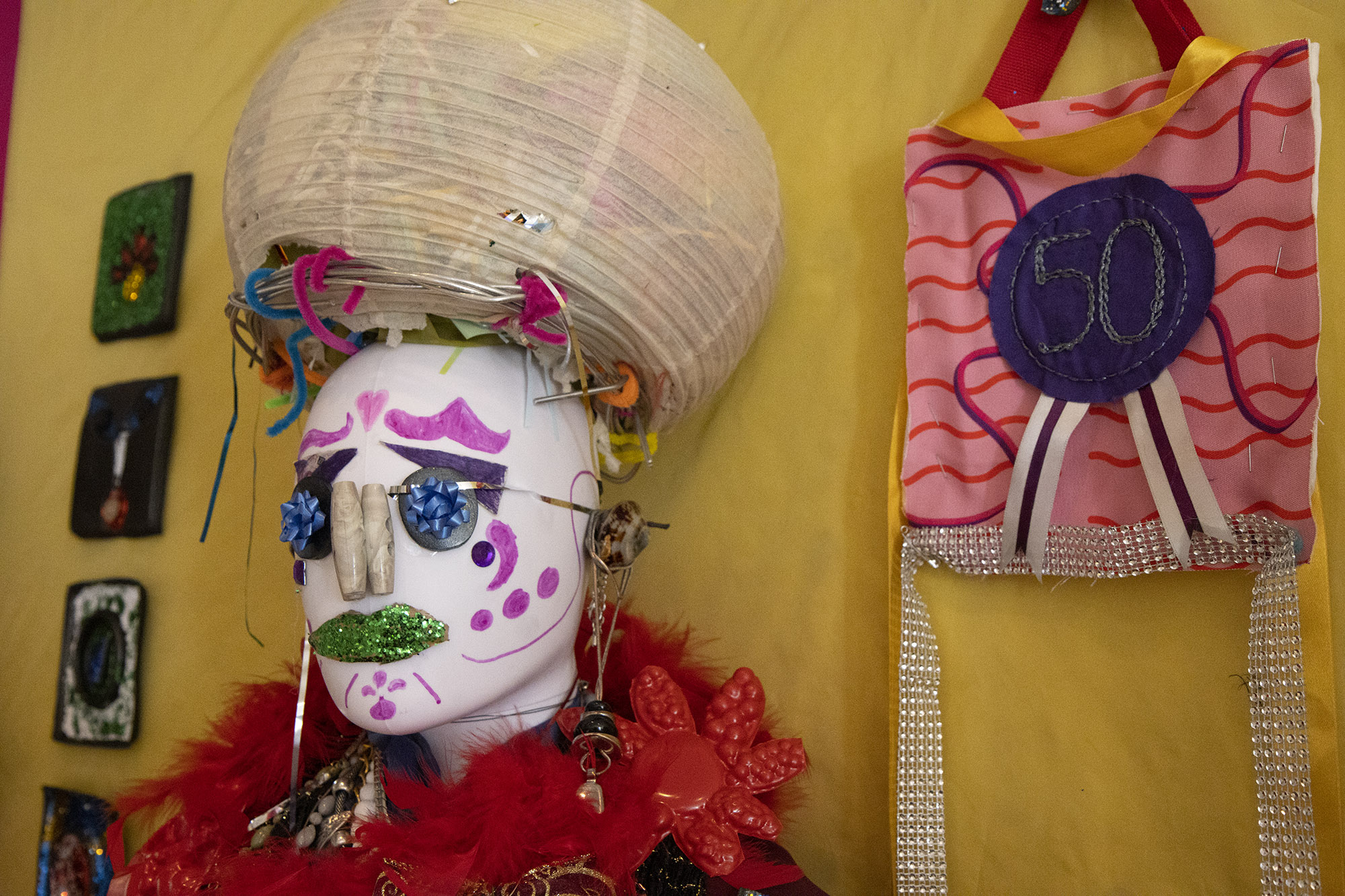 Close up of the head of a sculpture featuring a white mannequin face with painted features, glittering green lips, a button nose and ribbon bow eyes. There's a feather boa around its neck and it's wearing a headpiece made of a white paper pop-up lampshade stuffed with fabric and paper