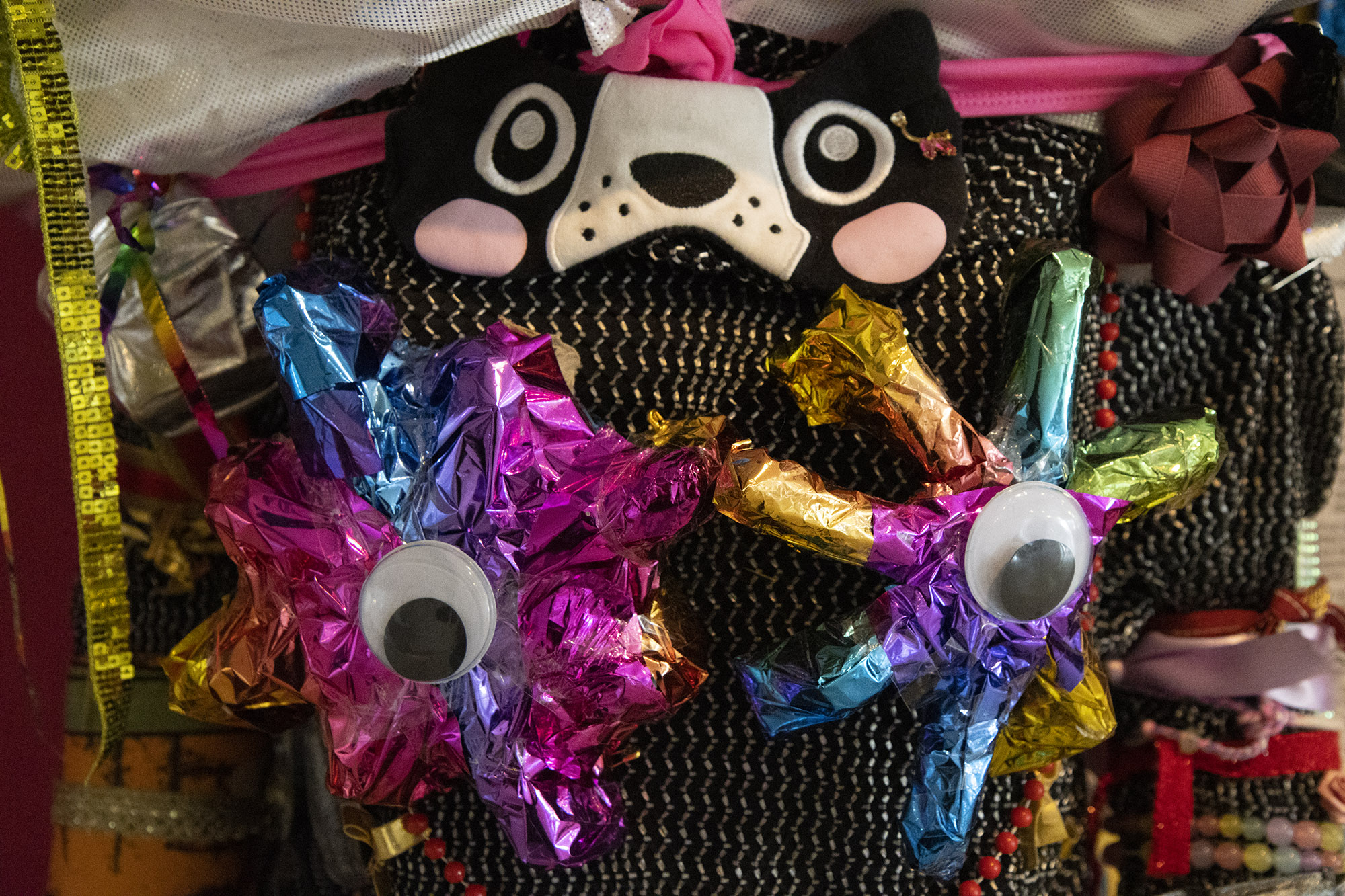 A close up of a section of sculpture with a black and white dog eye mask, a strip of gold, sequinned ribbon and two multicoloured foil stars with google eyes stuck to them
