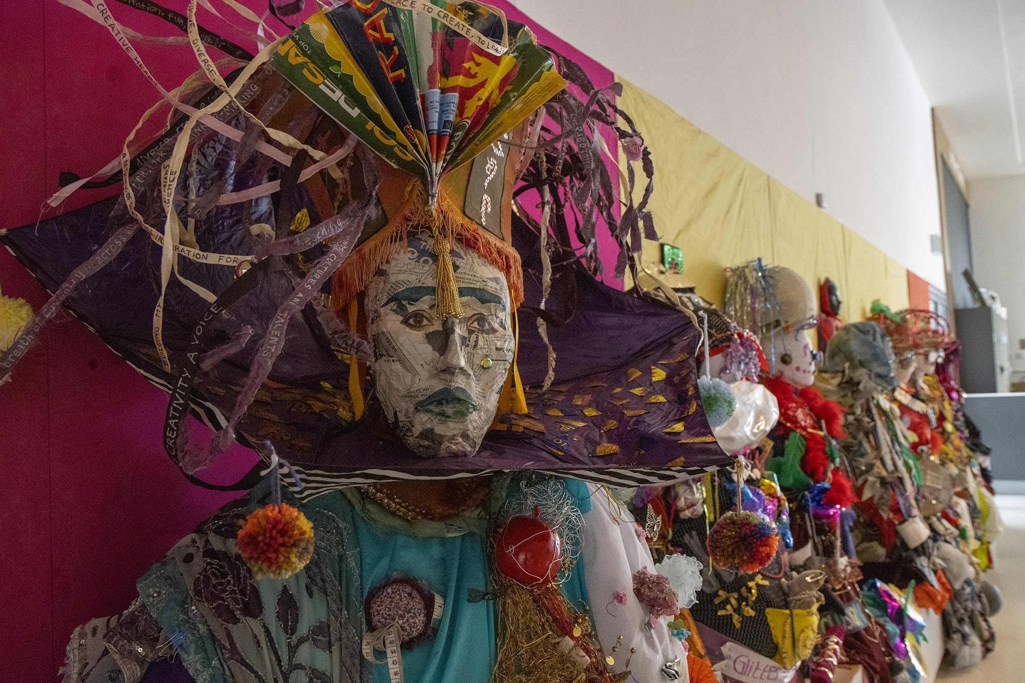 A set of sculptures on display in the Herbert. In the foreground is "Frida", a mannequin with a newspaper face and eyes cut out from a magazine. On top of her head is an orange, fringed lampshade with a coloured card fan fixed to the front, and ribbons with messages written on them. Around her neck is an upside down umbrella, painted with patterns and decorated with pom poms.