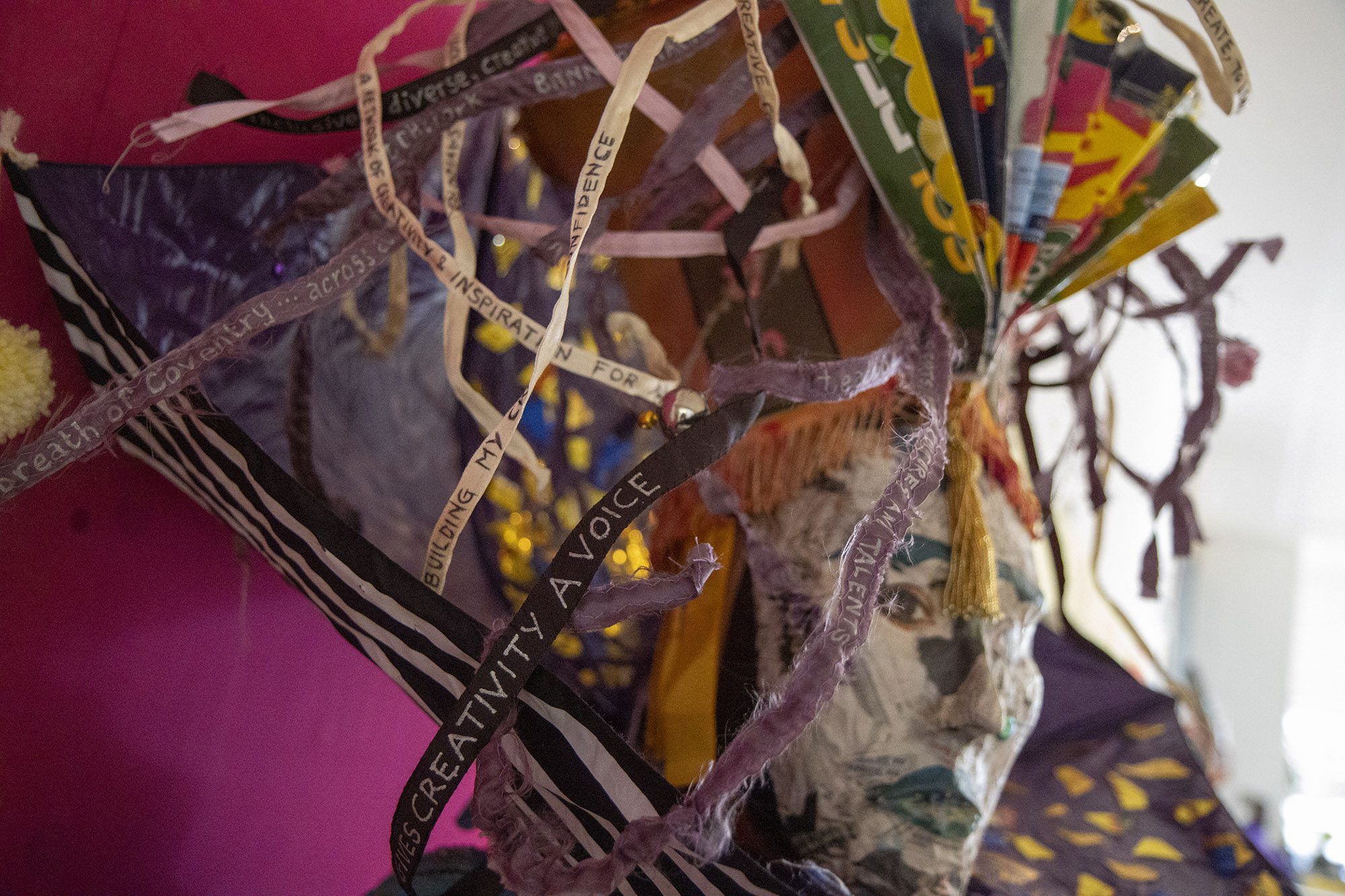 A close-up of "Frida", a mannequin with a newspaper face and eyes cut out from a magazine. On top of her head is an orange, fringed lampshade with a coloured card fan fixed to the front, and ribbons with messages written on them. Around her neck is an upside down umbrella, painted with patterns and decorated with pom poms.