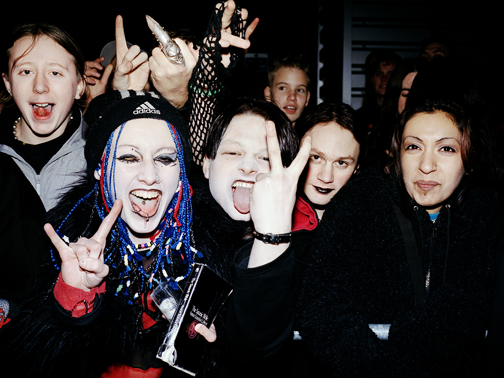 A group of young people in dark clothes and jewellery pulling funny faces and making hand gestures towards the camera. To the left of the frame, a young woman with white foundation, heavy eyeliner, bright blue braids and lip and tongue piercings is holding a copy of The Satanic Bible. Other members of the group are wearing, variously, black lipstick, a fishnet glove and a patterned metal finger covering. 