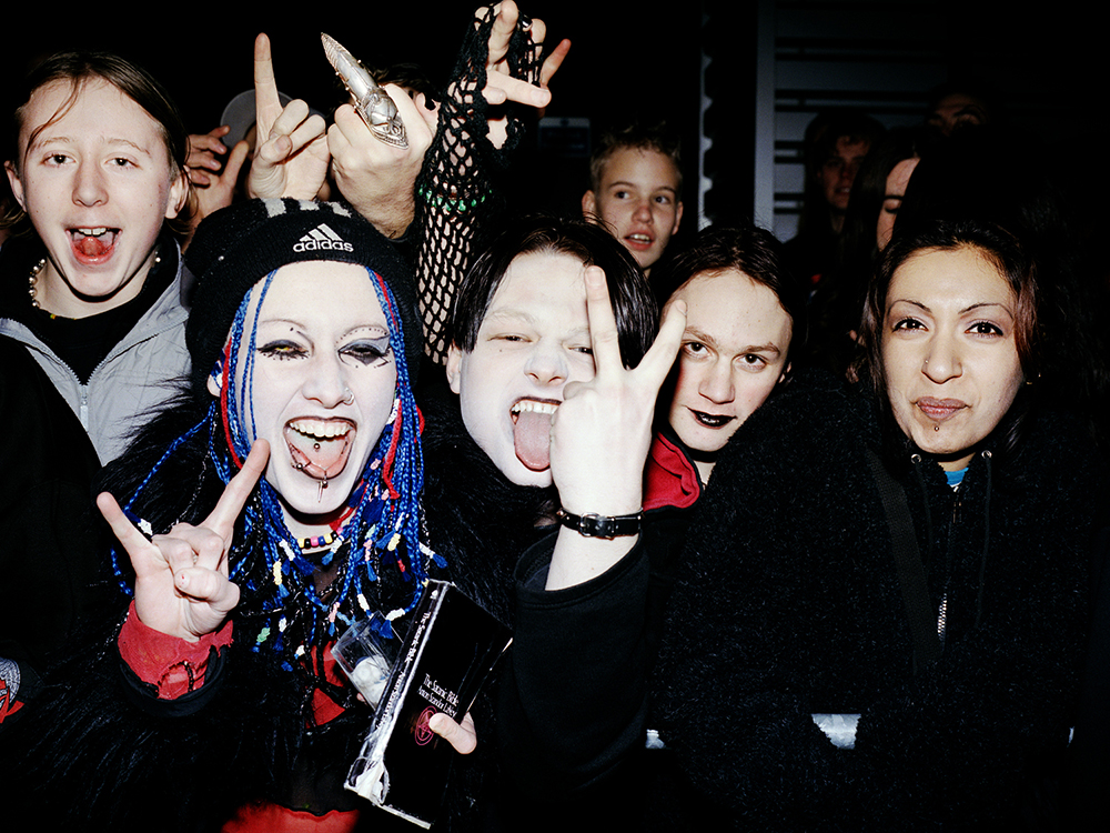 A group of young people in dark clothes and jewellery pulling funny faces and making hand gestures towards the camera. To the left of the frame, a young woman with white foundation, heavy eyeliner, bright blue braids and lip and tongue piercings is holding a copy of The Satanic Bible. Other members of the group are wearing, variously, black lipstick, a fishnet glove and a patterned metal finger covering.