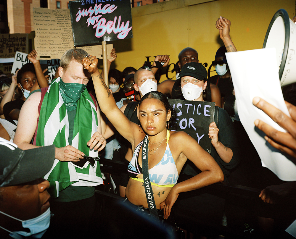 A photo of a Black Lives Matter protest. At the front of the group, a young woman in a bikini top and hooped earrings is raising her fist in the air. Beside her, a young man in a green spotted gaiter has a green and white flag draped around his neck. Behind them, a group of mostly masked people are gathered, holding up signs with the names of people murdered. Just in view to the right of the frame is part of a megaphone.