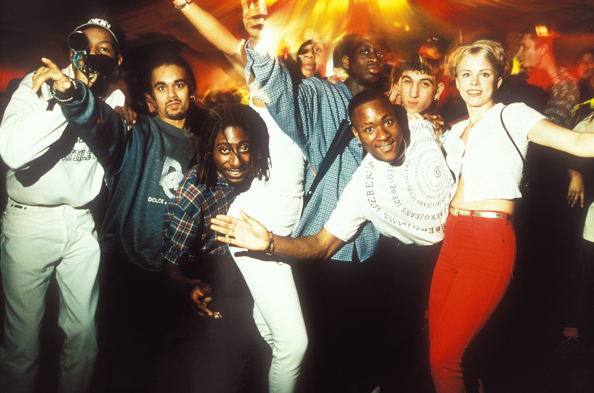 A group of young men and women of different ethnicities posing at what appears to be a rave, with bright red and yellow light pouring down and people dancing behind them.
