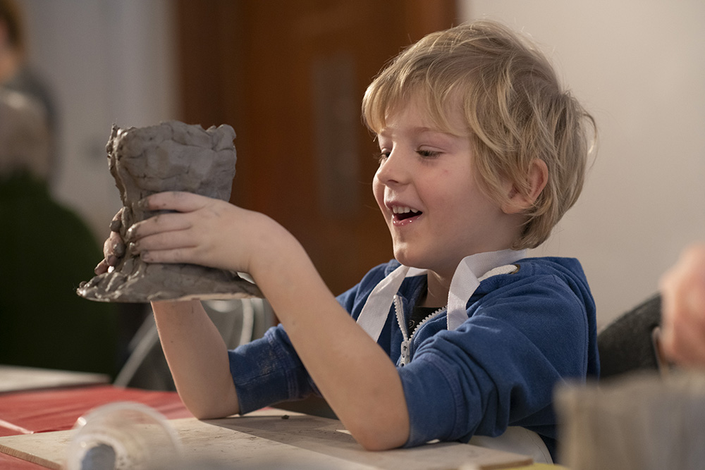 A photograph of a smiling boy sculpting a shape out of clay