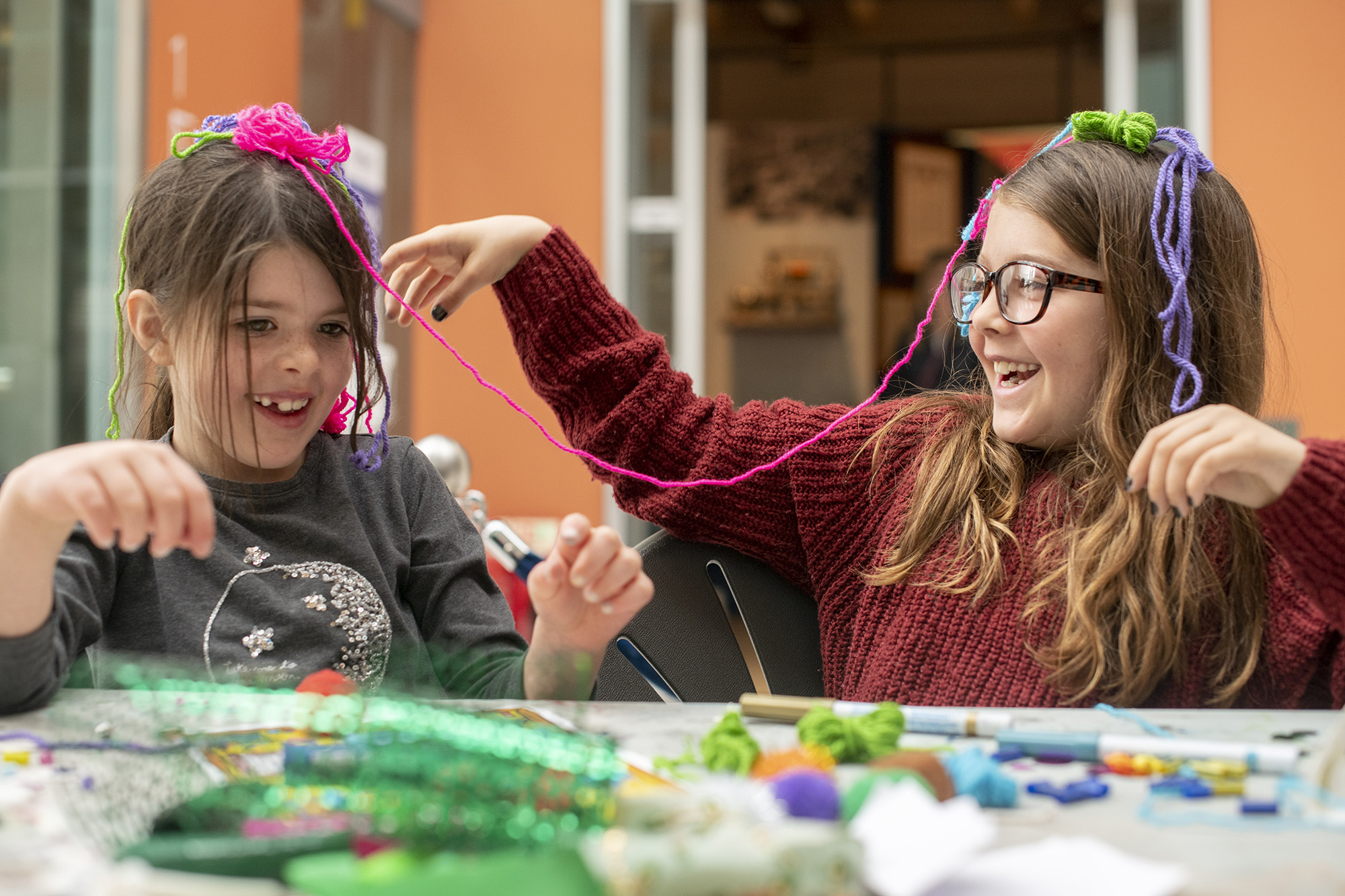 Two girls sitting at a craft table, playing with coloured yarn that is stretched across their heads