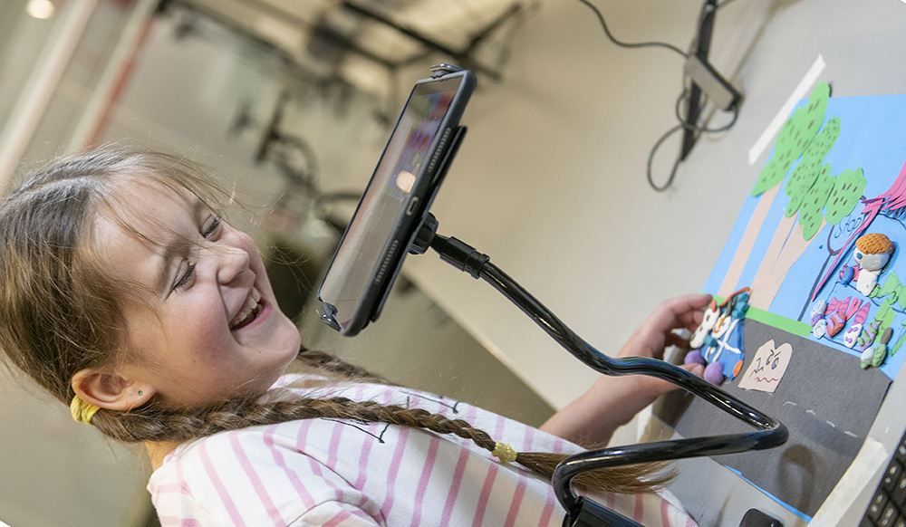 A laughing girl working on a stop-motion animation, using an electronic tablet to take photos of a scene composed of a paper backdrop and plasticine characters