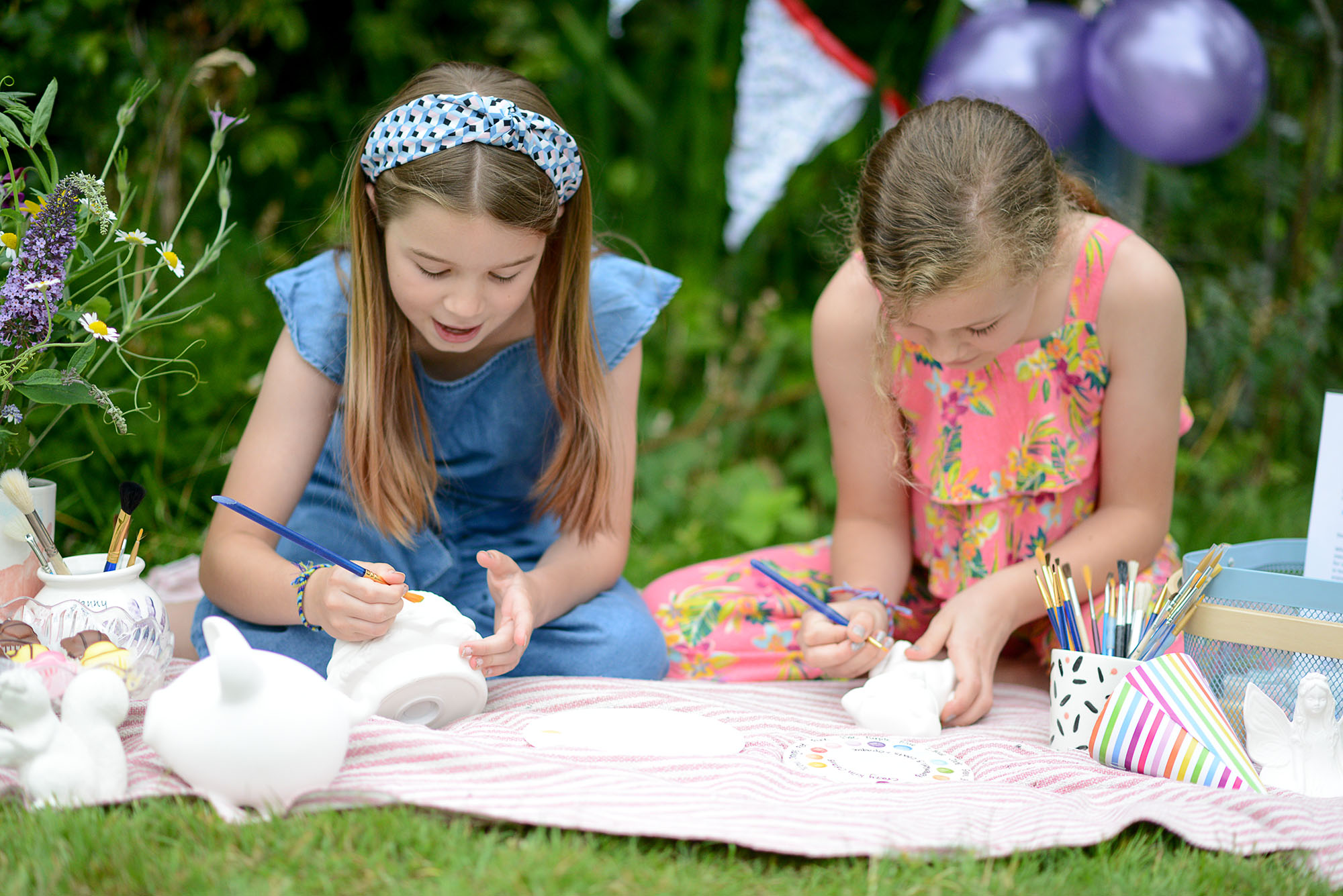 Two girls sitting outside on a picnic blanket painting white ceramic pots