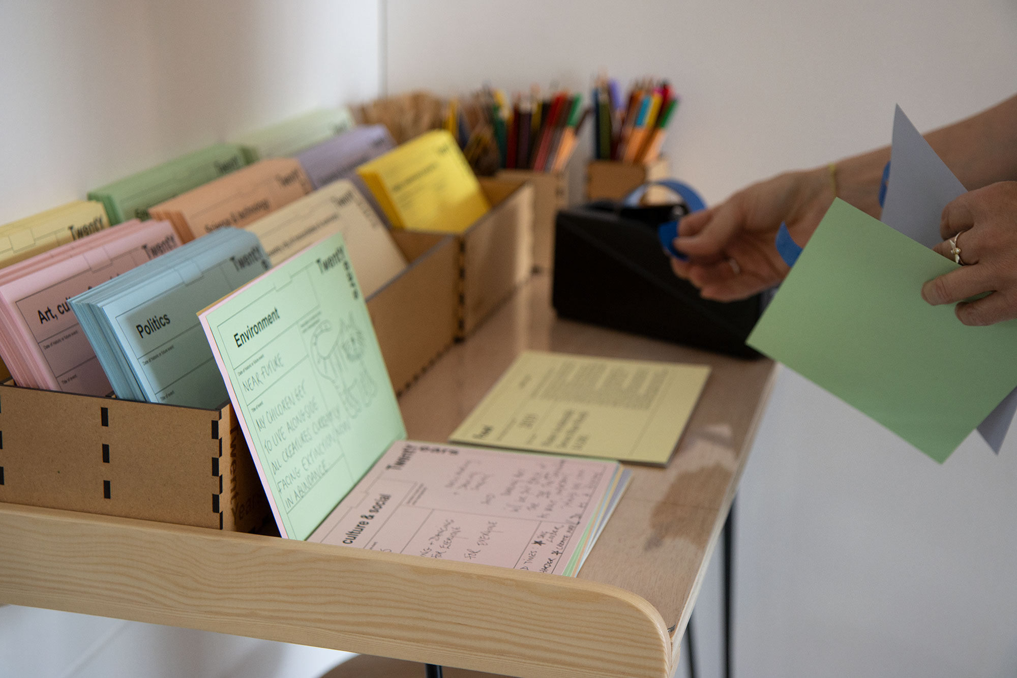 A table laid with coloured notecards, pens and pencils and a tape dispenser. A hand is pulling tape from the dispenser while another holds two of the notecards.