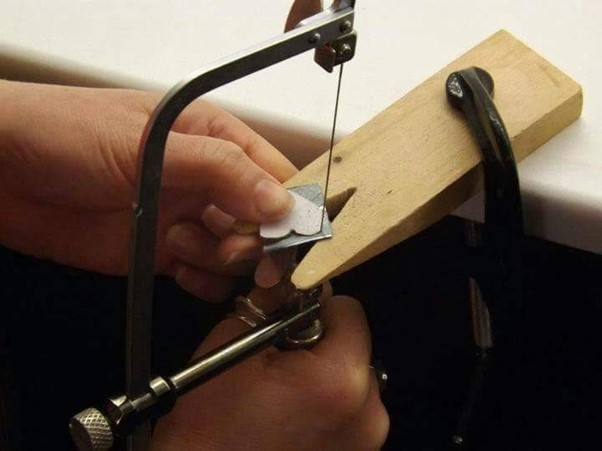 A close up of hands and a hacksaw, cutting a heart shape out of a small square of silver resting against a wooden block