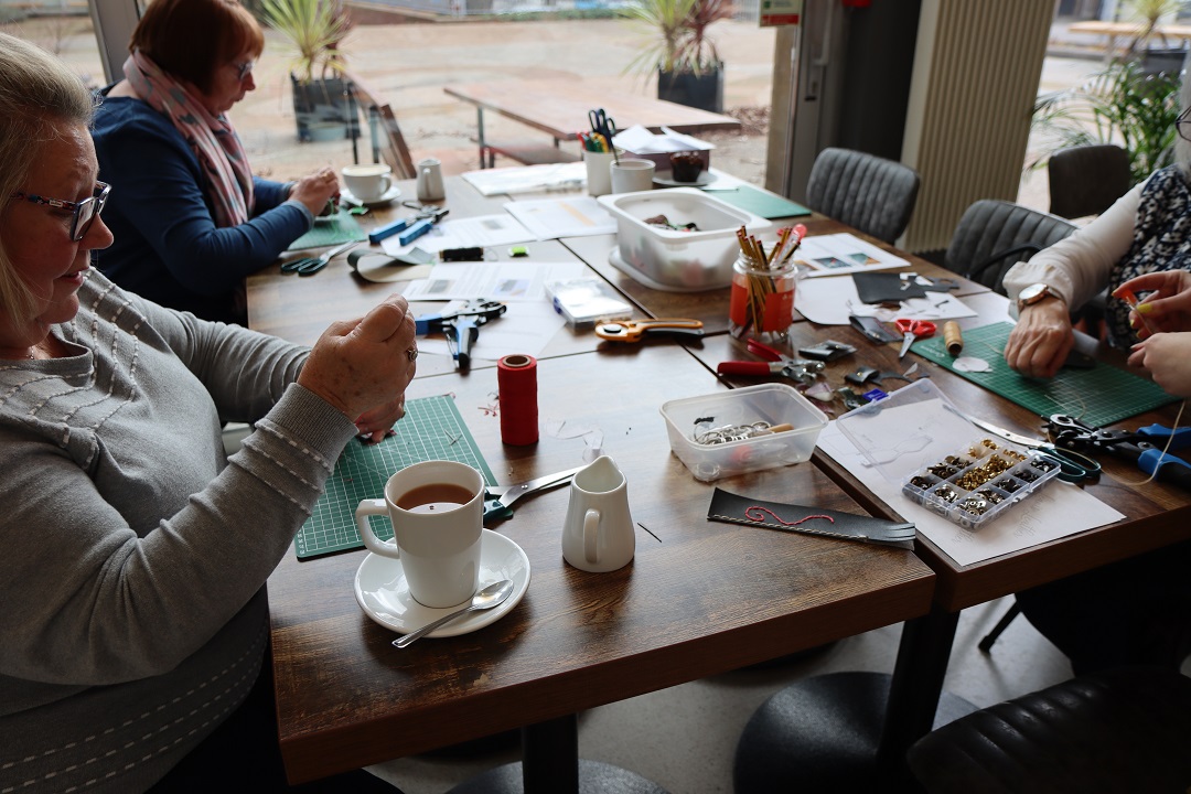 A group of people sitting around a table in Alfred's Cafe, working on various craft projects with hot drinks beside them