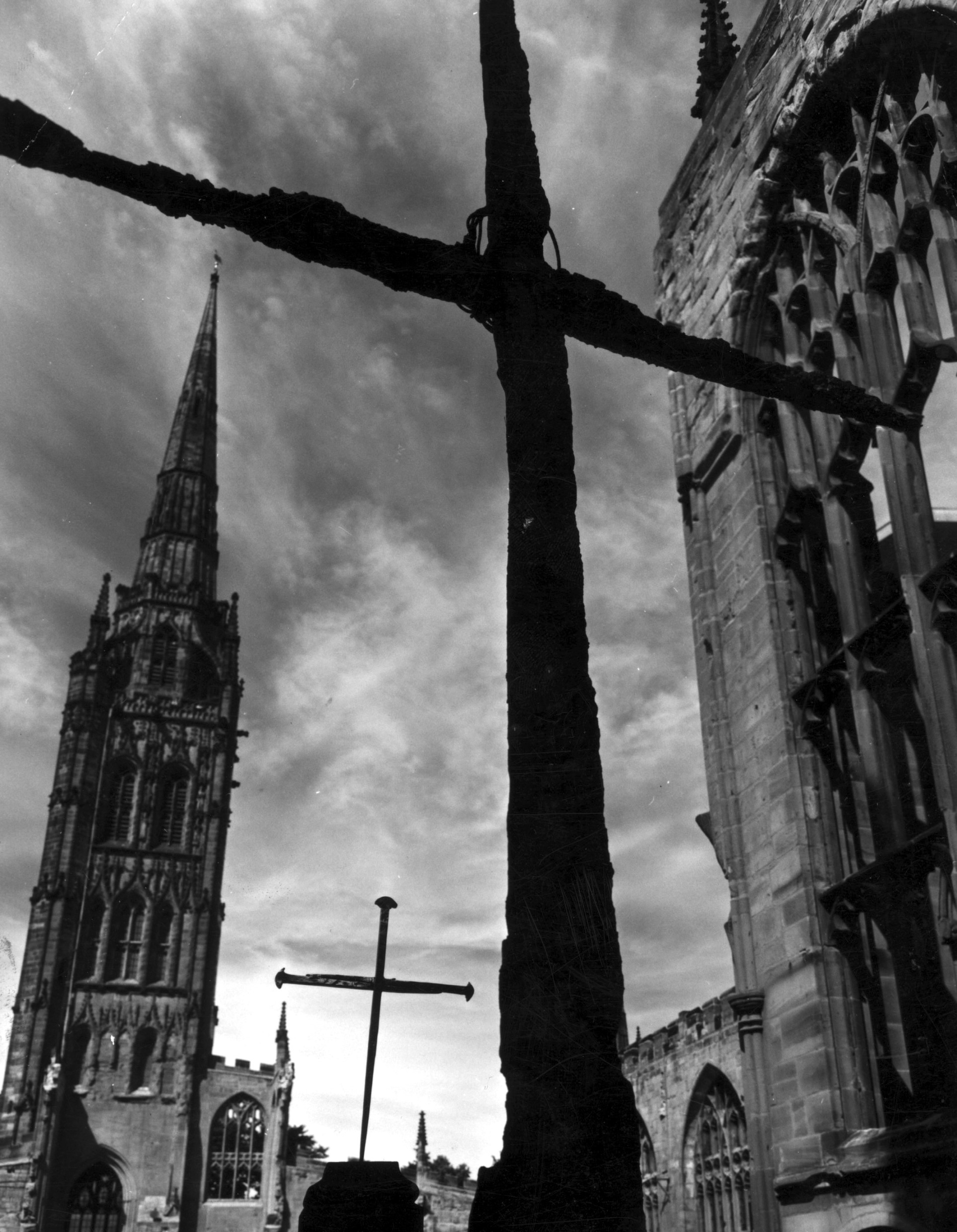 A black and white photograph taken in Coventry Cathedral Ruins, with the burned cross on the altar silhouetted in the foreground against a dramatic sky and the top of one of the windows. In the background is the old spire and a large-scale recreation of the cross of nails
