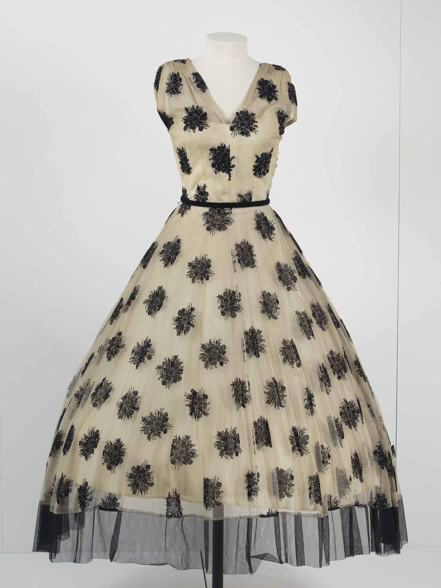 A formal 1950s v-necked, fit and flare dress in cream with a black floral pattern, short sleeves and a cinched black waistband