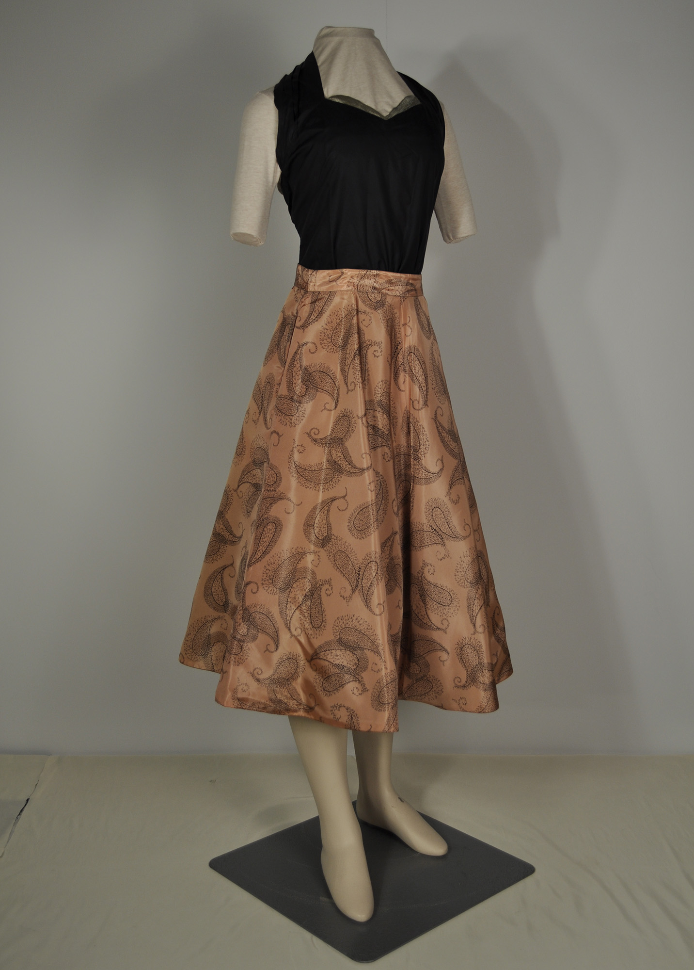 A 1950s woman's outfit consisting of a sleeveless, fitted black top with a sweetheart neckline and a peach coloured circle skirt with a paisley print
