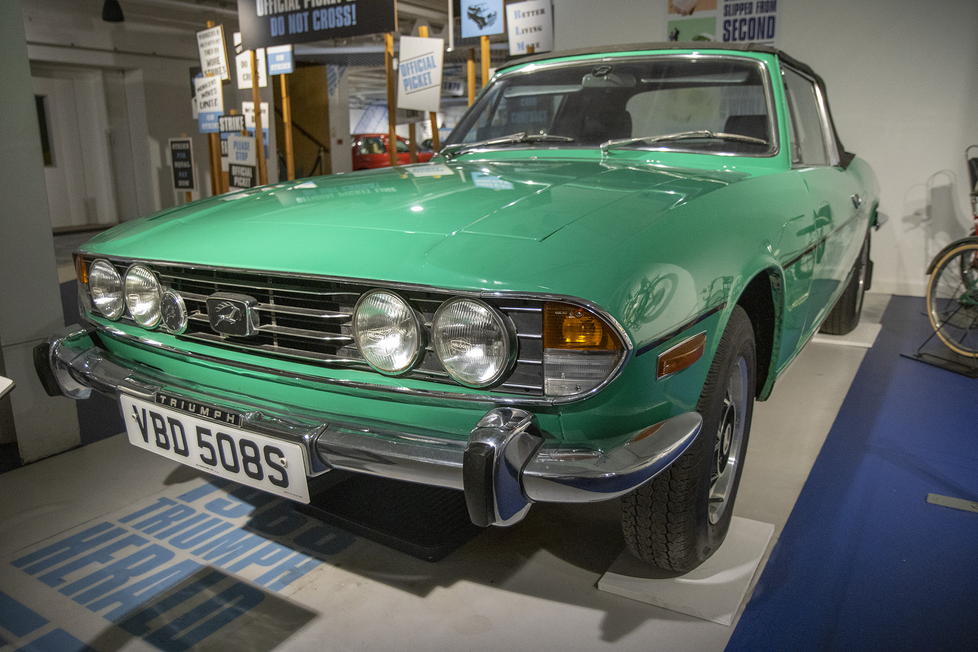 A green Triumph Stag on display in Coventry Transport Museum, in front of a model picket line that visitors can walk through.