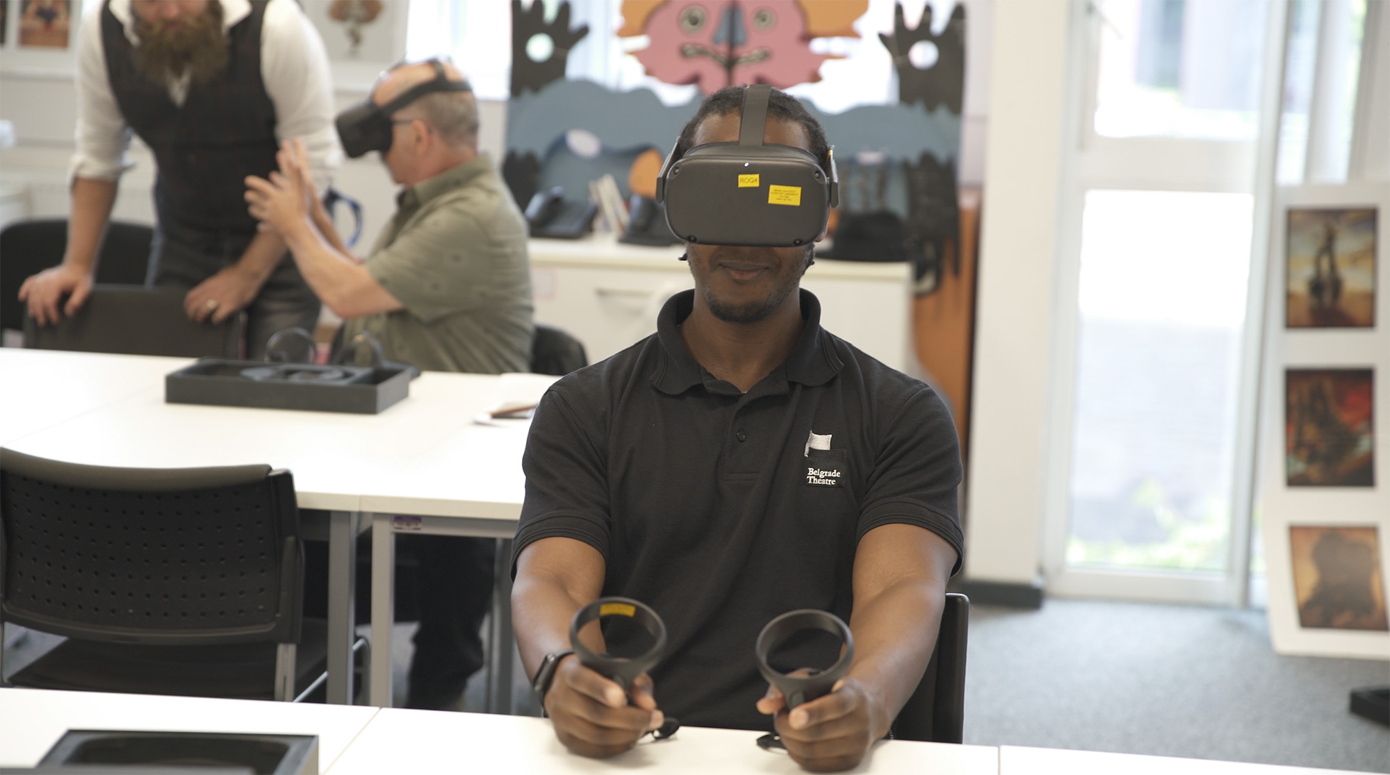 A man sitting at a table wearing a VR headset and using a VR handset. In the background are two other people talking