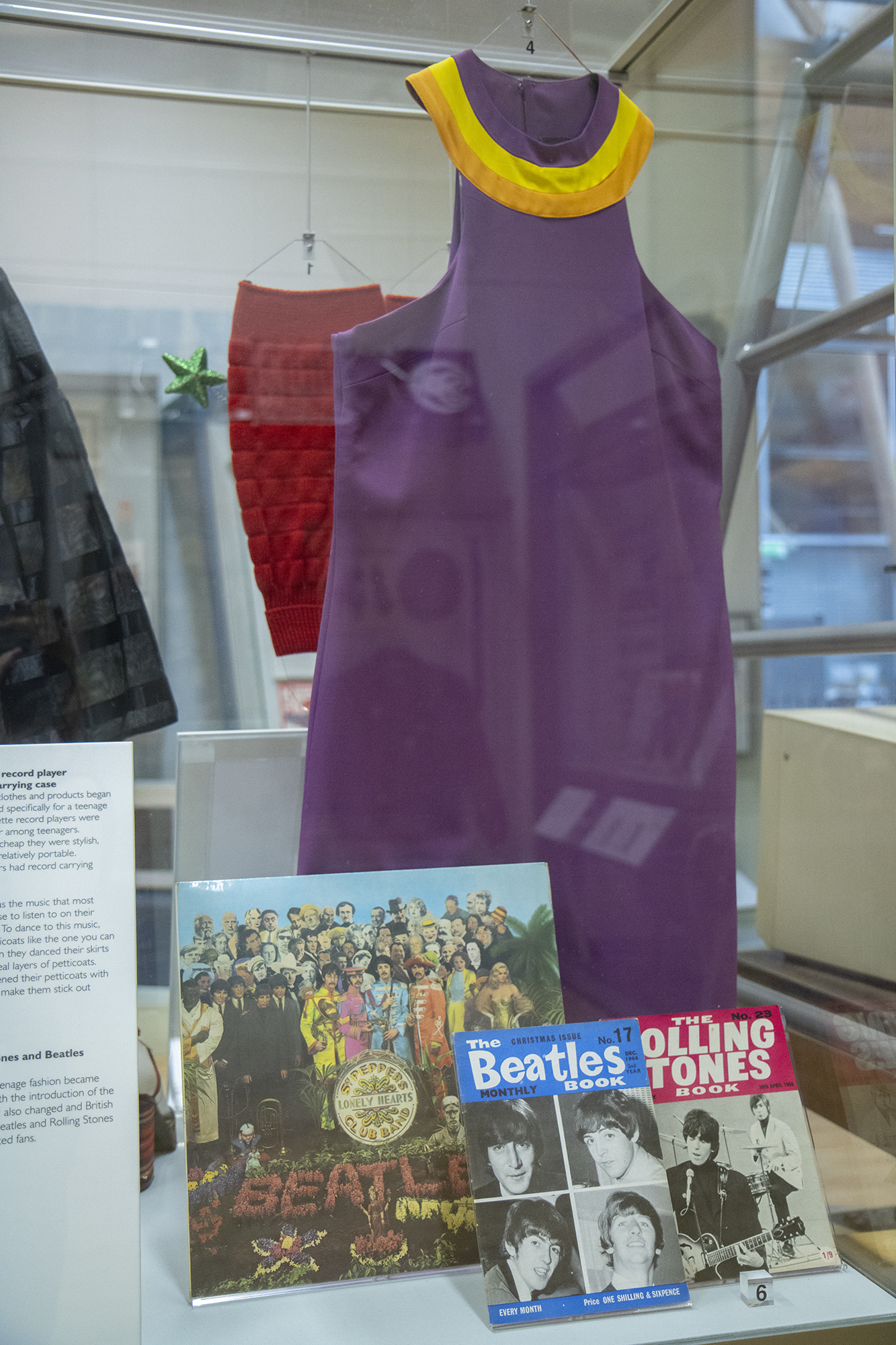 A photograph of a purple, sleeveless mini dress with a bright yellow and orange collar on display in the Herbert's History Gallery alongside a Sgt Pepper's Lonely Hearts Club Band record and two fanzines - one dedicated to the Beatles and the other to the Rolling Stones