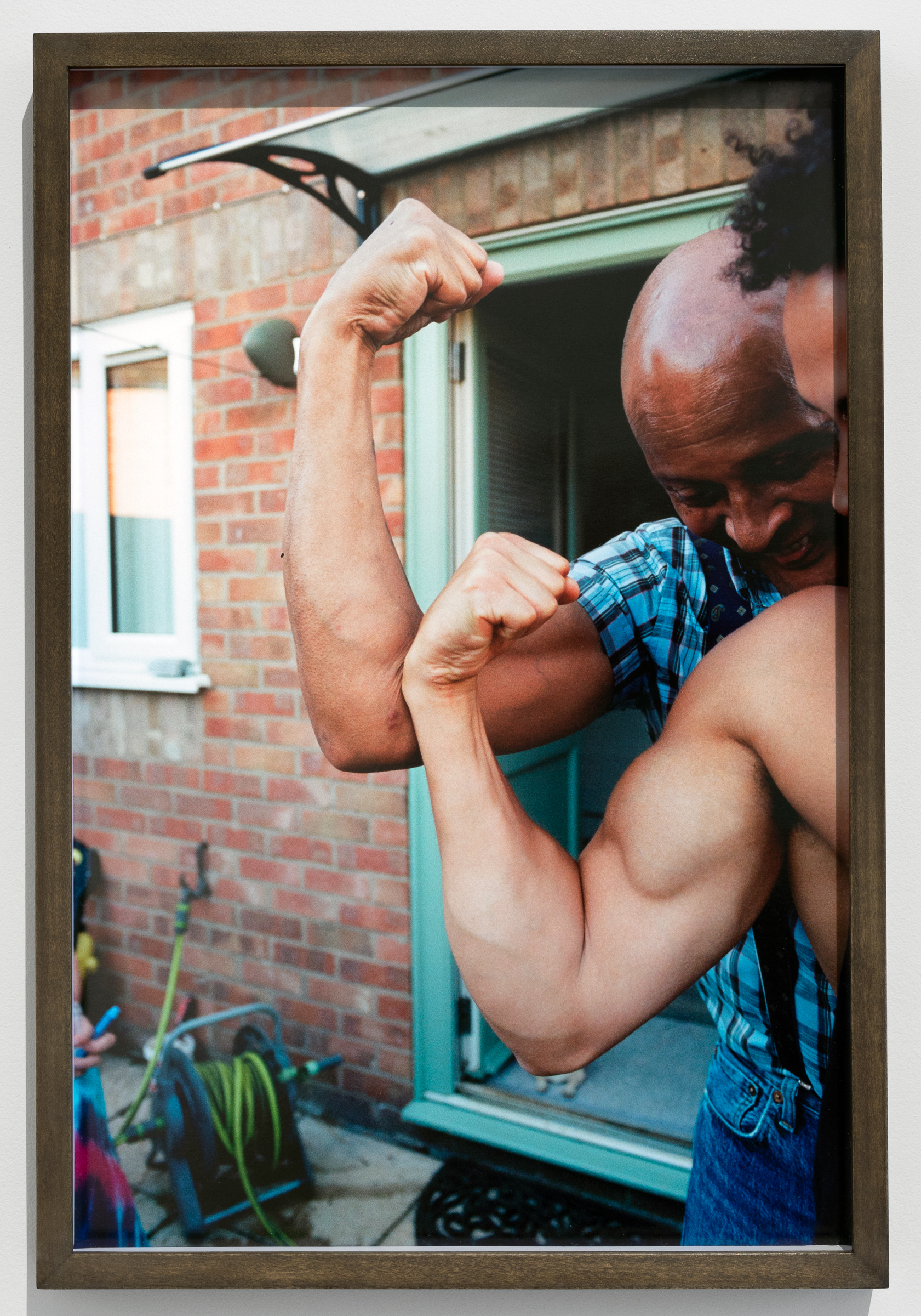 Rene Matić's Paul and Zac in the Garden, Peterborough (2022) - a photograph of two men flexing their biceps outside, in front of an open door. 