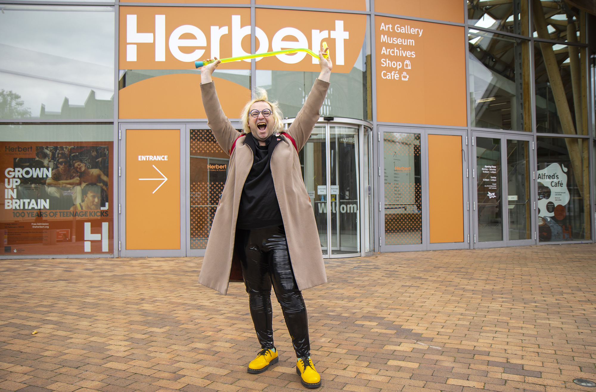 Alex Billingham stands outside the Herbert Art Gallery & Museum in a defiant pose: mouth open and arms raising a neon yellow perspex walking stick high in the air. She is wearing a black top, black PVC trousers, bright yellow shoes and a light tan coloured coat.