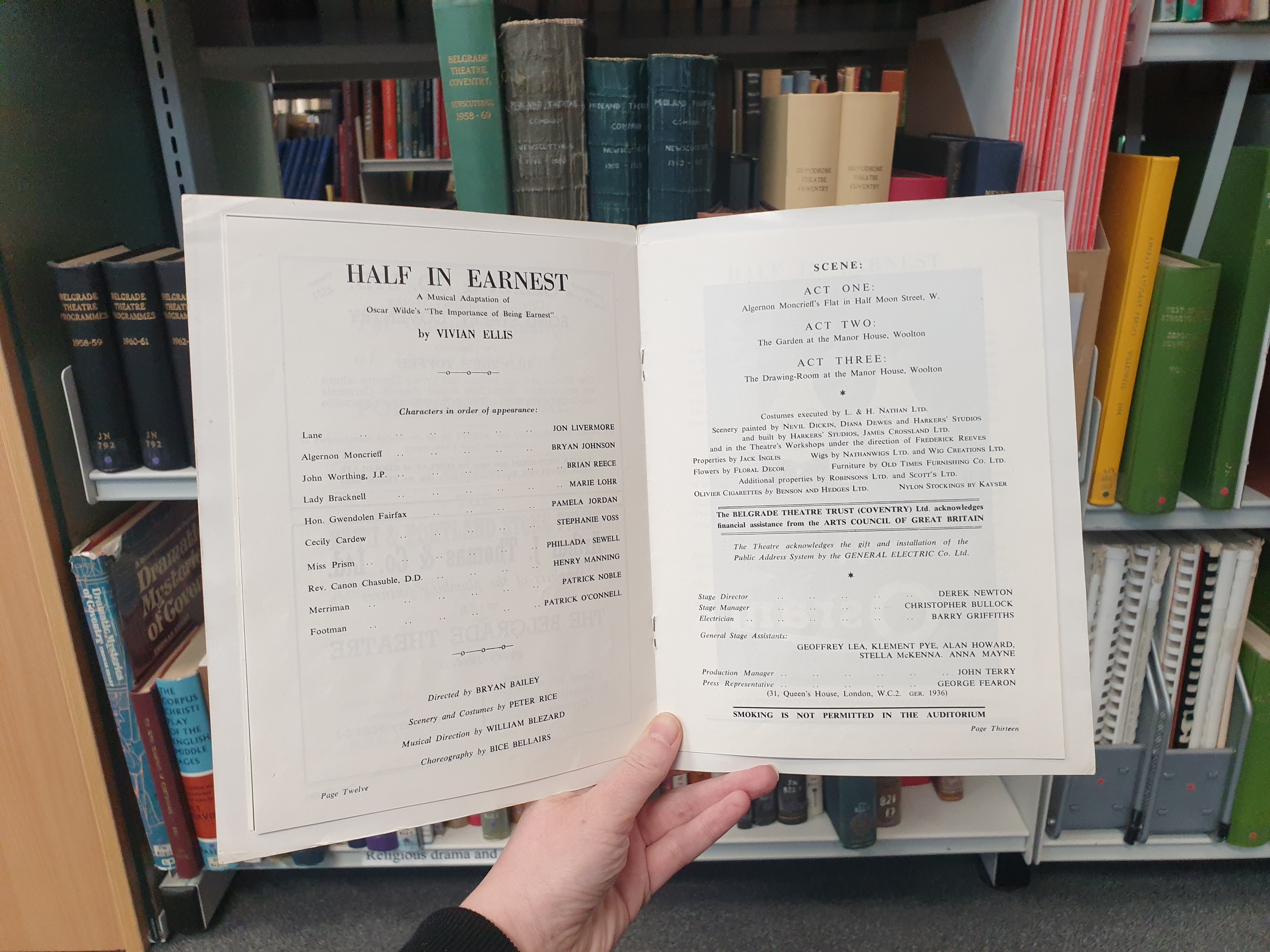 An open programme for Half in Earnest at the Belgrade Theatre featuring a list of cast and creatives and a scene breakdown. The programme is being held up in front of a bookcase in Coventry Archives