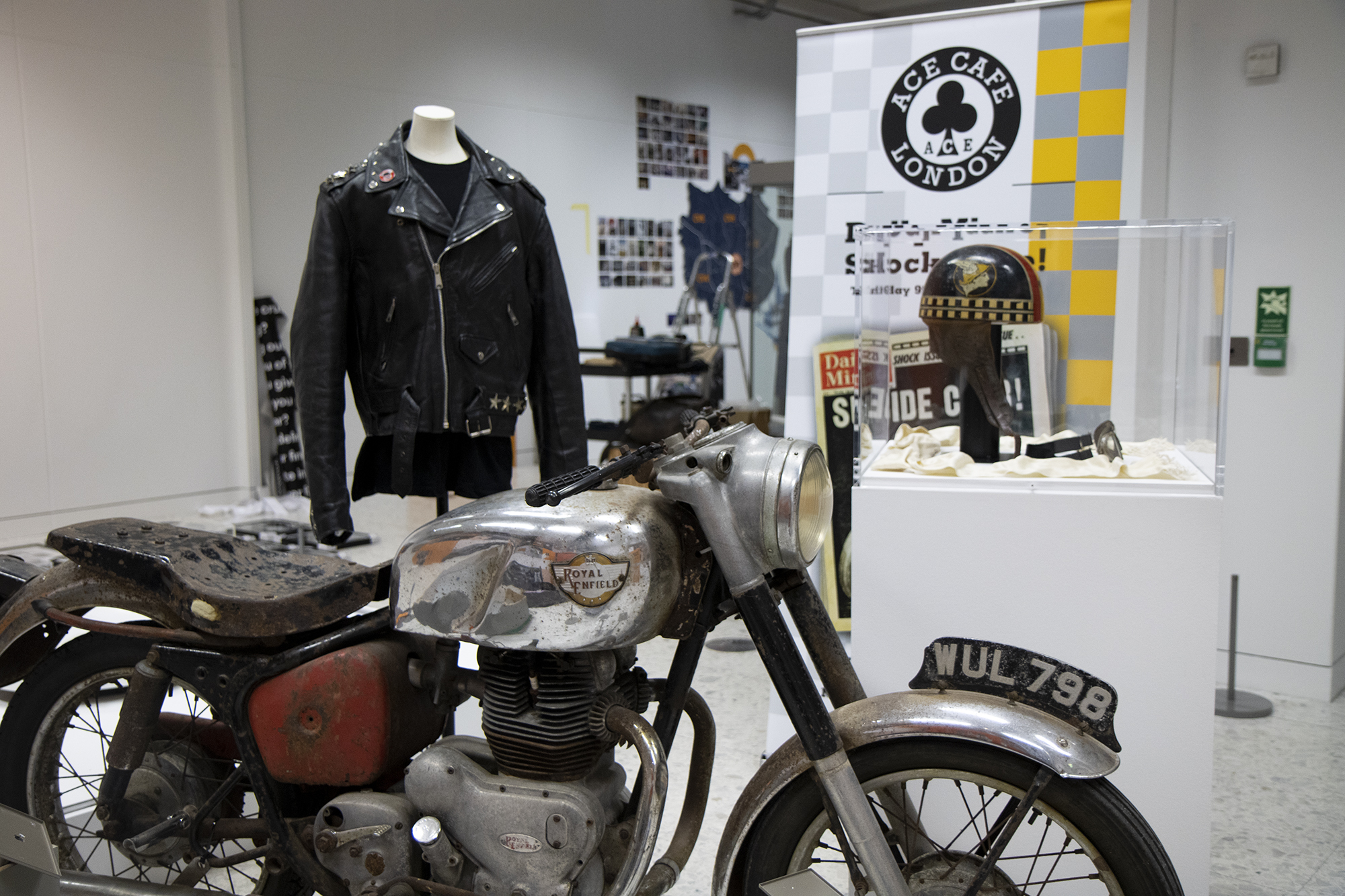 A photograph of a leather jacket, Royal Enfield motorcycle, helmet, goggles and a pop-up banner featuring the Ace Cafe logo, all on display in Grown Up in Britain