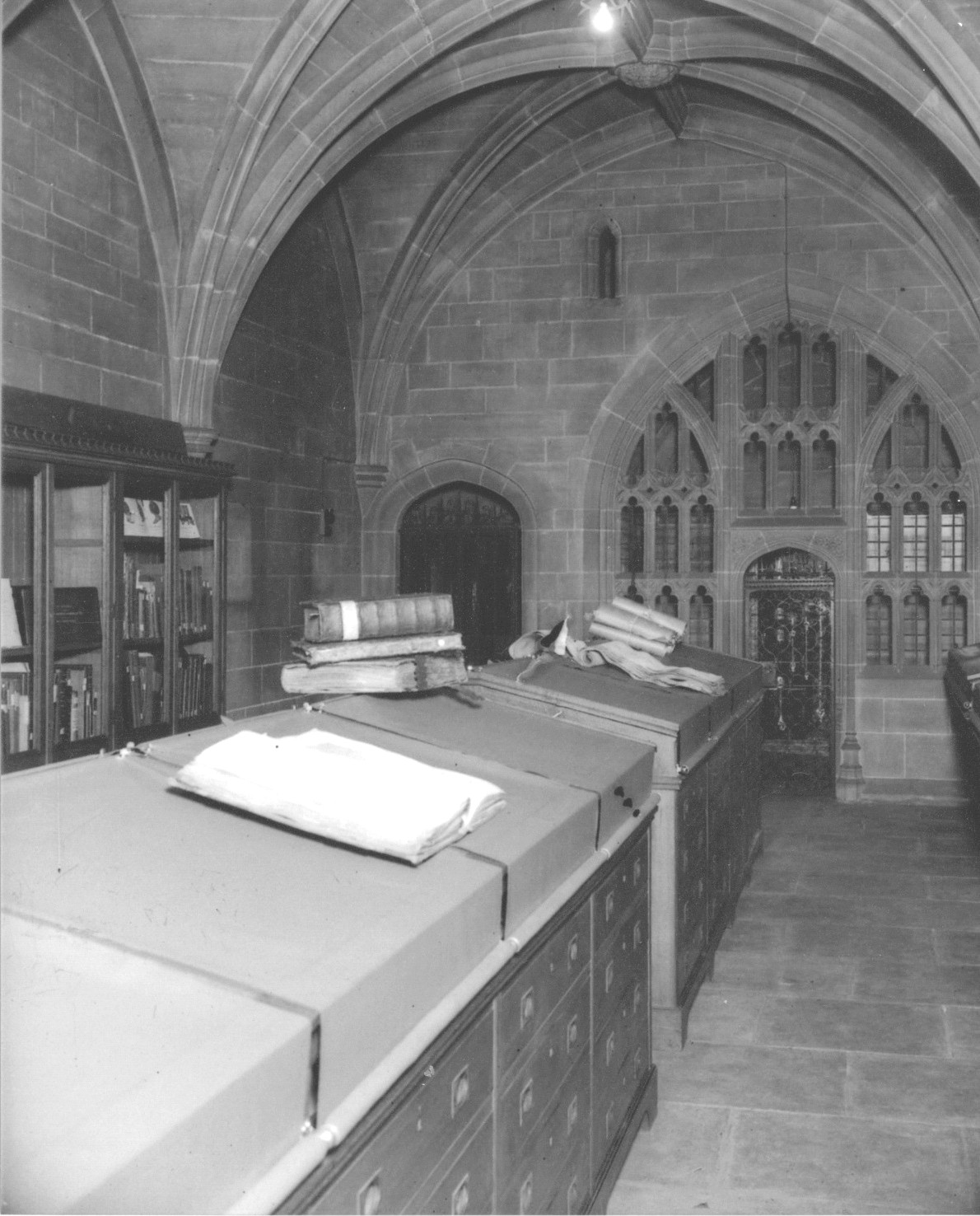 A black and white photograph of the inside of the Muniment Room at St Mary's Guildhall - a medieval building with an arched ceiling and gothic windows. Inside the room are books resting on top of a filing cabinet.