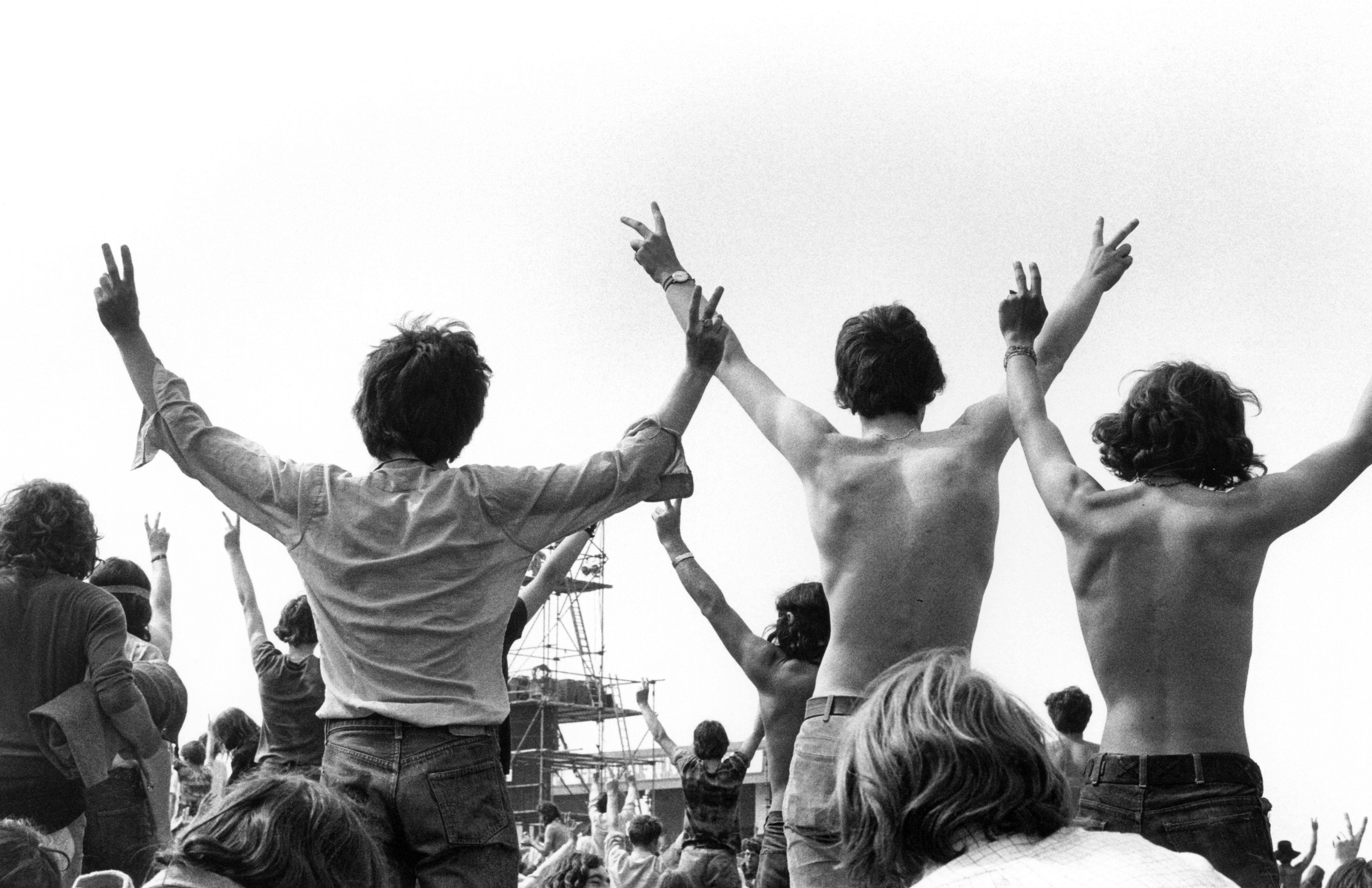 A crowd of people at a festival - we can see their backs as they face towards the stage. Some young men are sitting on the shoulders of others, making peace signs with their hands