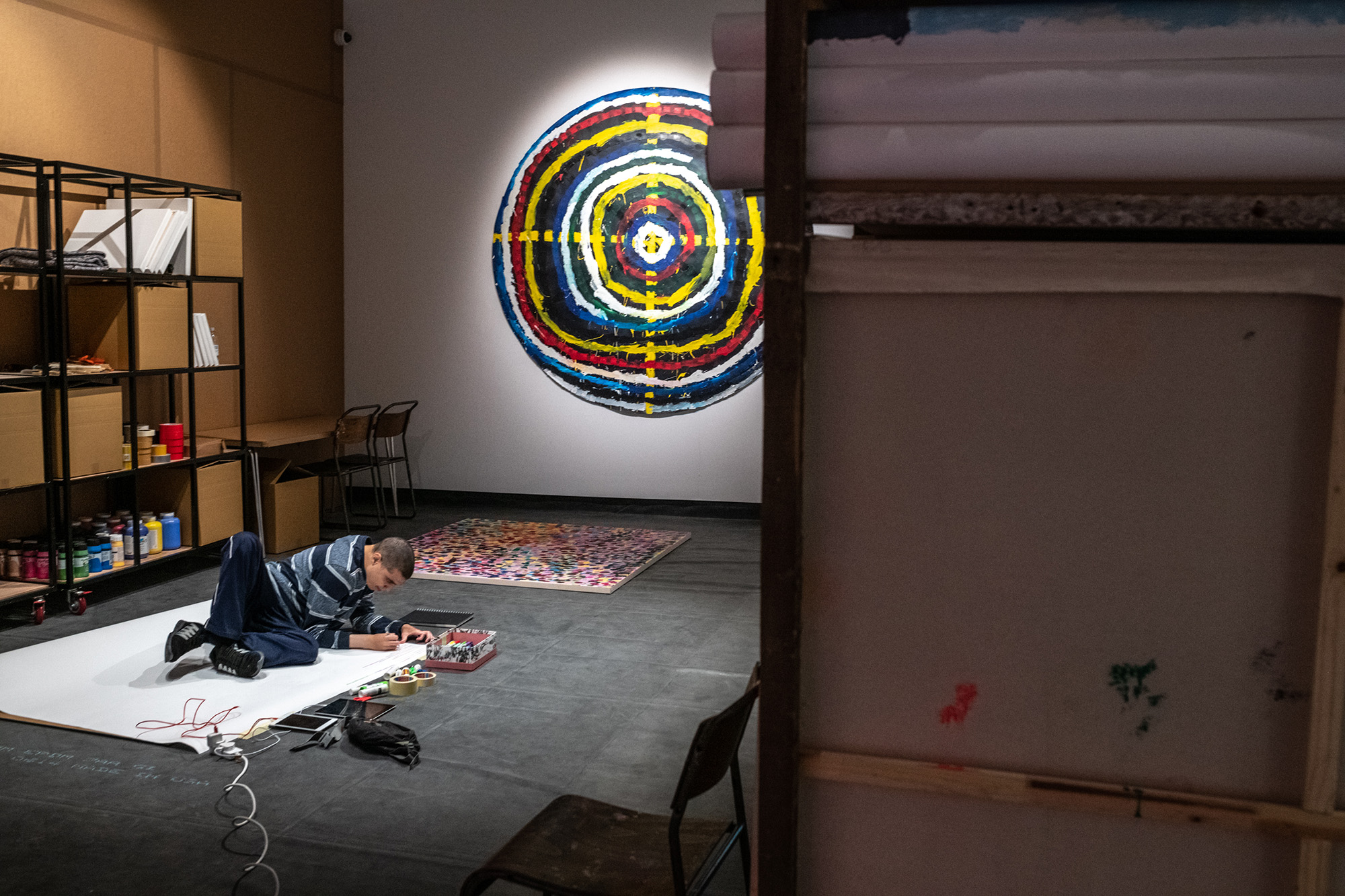 A photograph from the Project Art Works section of the Turner Prize exhibition at the Herbert Art Gallery and Museum. The image shows a young man leaning on a large sheet of paper on the floor, about to draw or write on the paper. Around him are pens, rolls of tape, ipads, a charger and a tin containing additional stationery. Large storage units with cardboard trays and shelves in a metal frame stand tall behind him. Hanging on one wall is a large, circular artwork, consisting of rings painted in different colours, like a target.